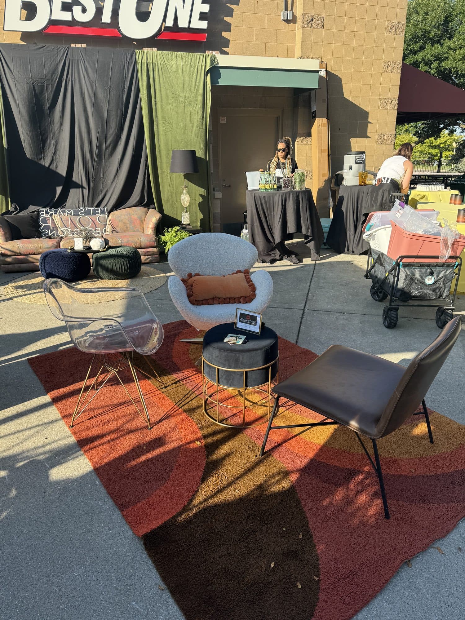 The lounge staged on a painted concrete lot, ghost chairs and a clay accent pillow around a gold side table, beneath a BestOne building sign.