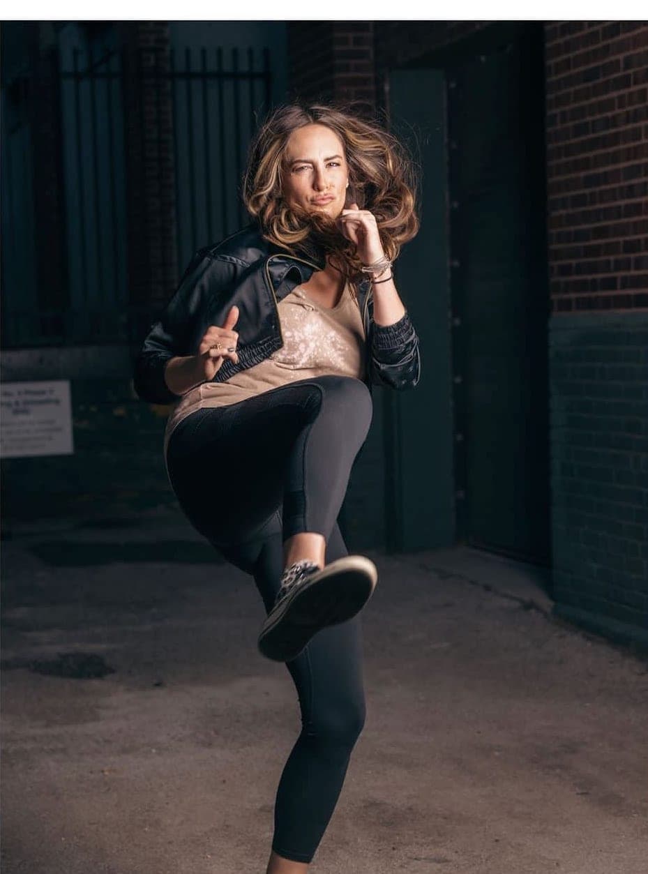 Haley mid high-kick in an alley, leather jacket and sequined tank, hair flying, brick wall and iron gate behind her.