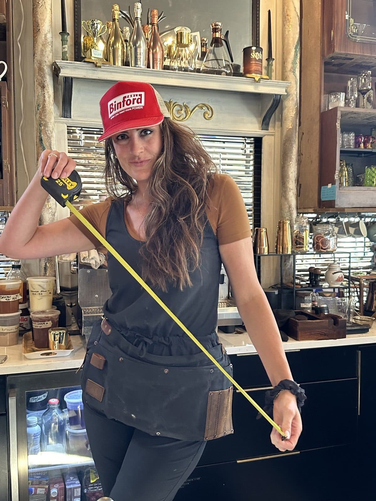 Haley in a red cap and apron pulling a tape measure in a workshop full of tools and paint.