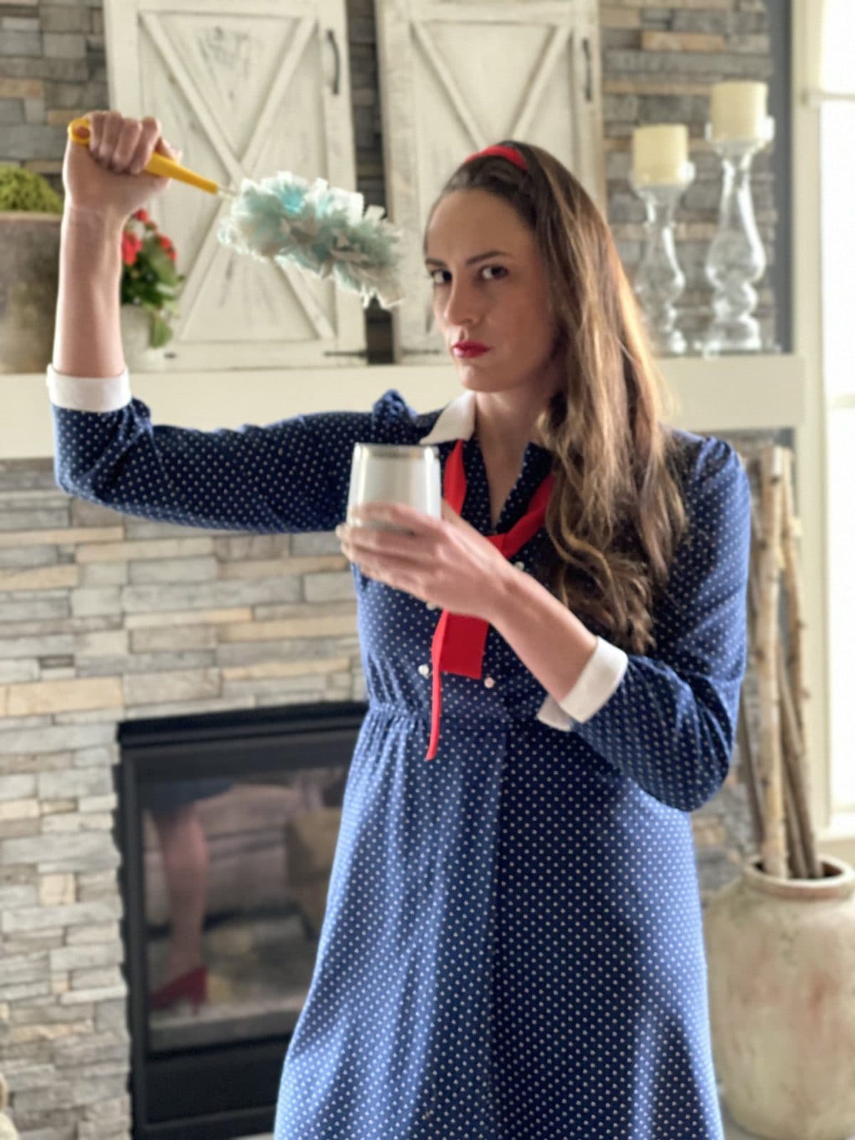 Haley in a navy polka-dot dress with a feather duster and a jar of fresh flowers in front of a stone fireplace.