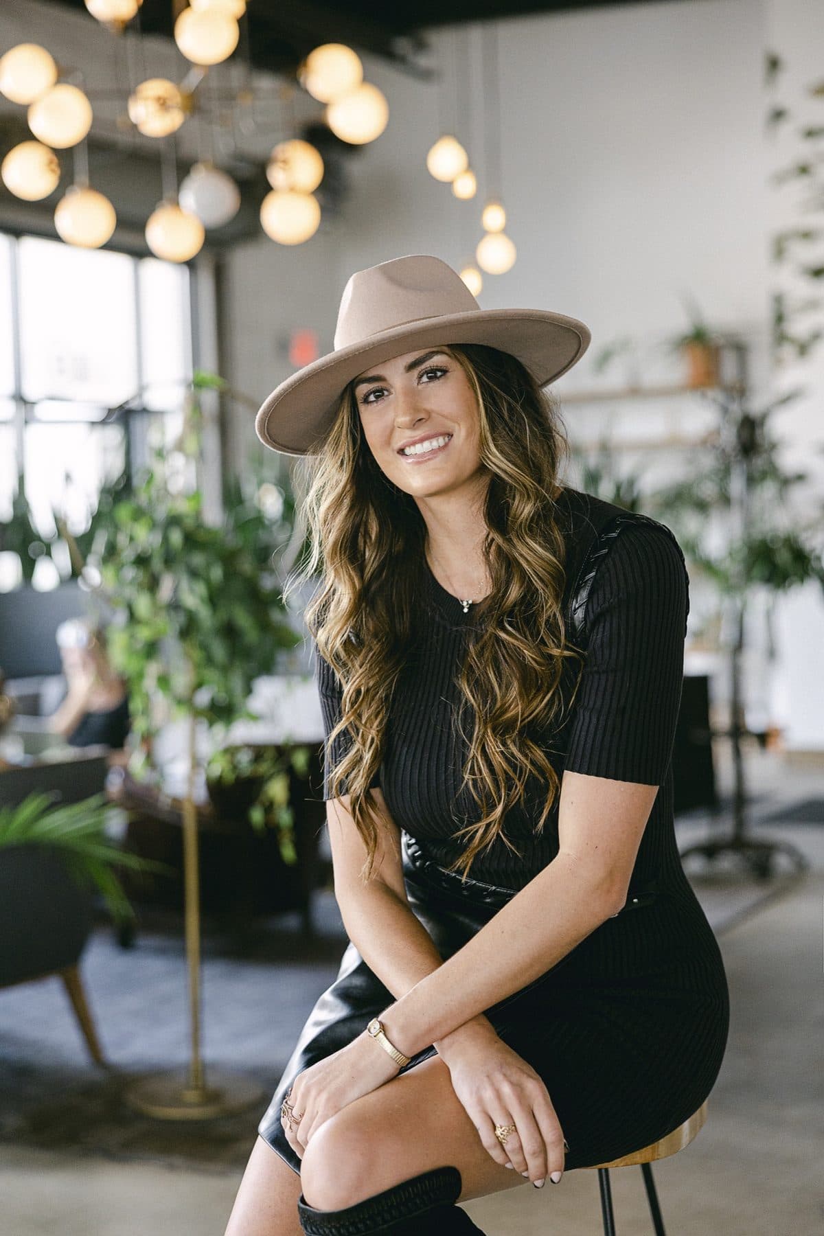 Perched on a stool in a black knit dress and cream fedora, string lights behind.