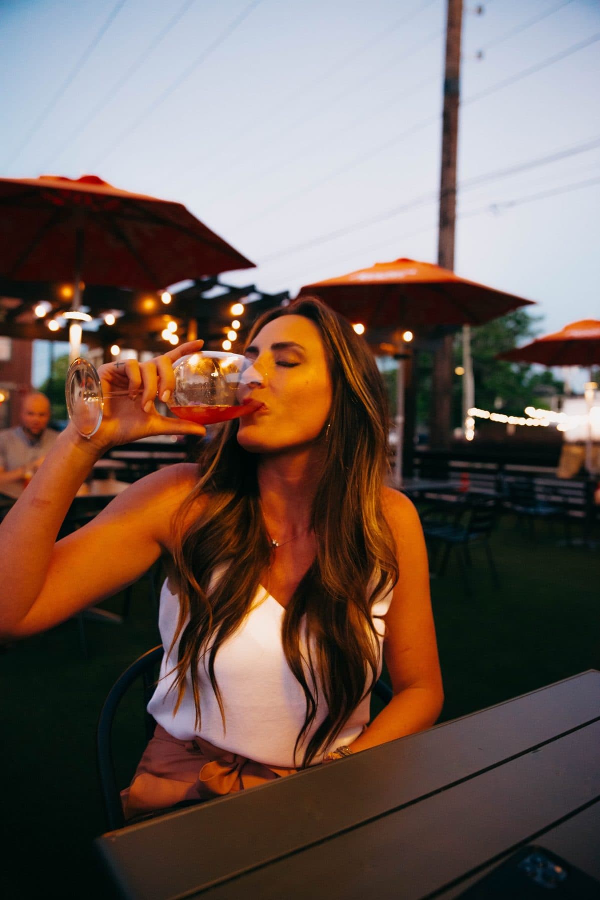 Sipping a drink on a patio under red umbrellas, white tank.