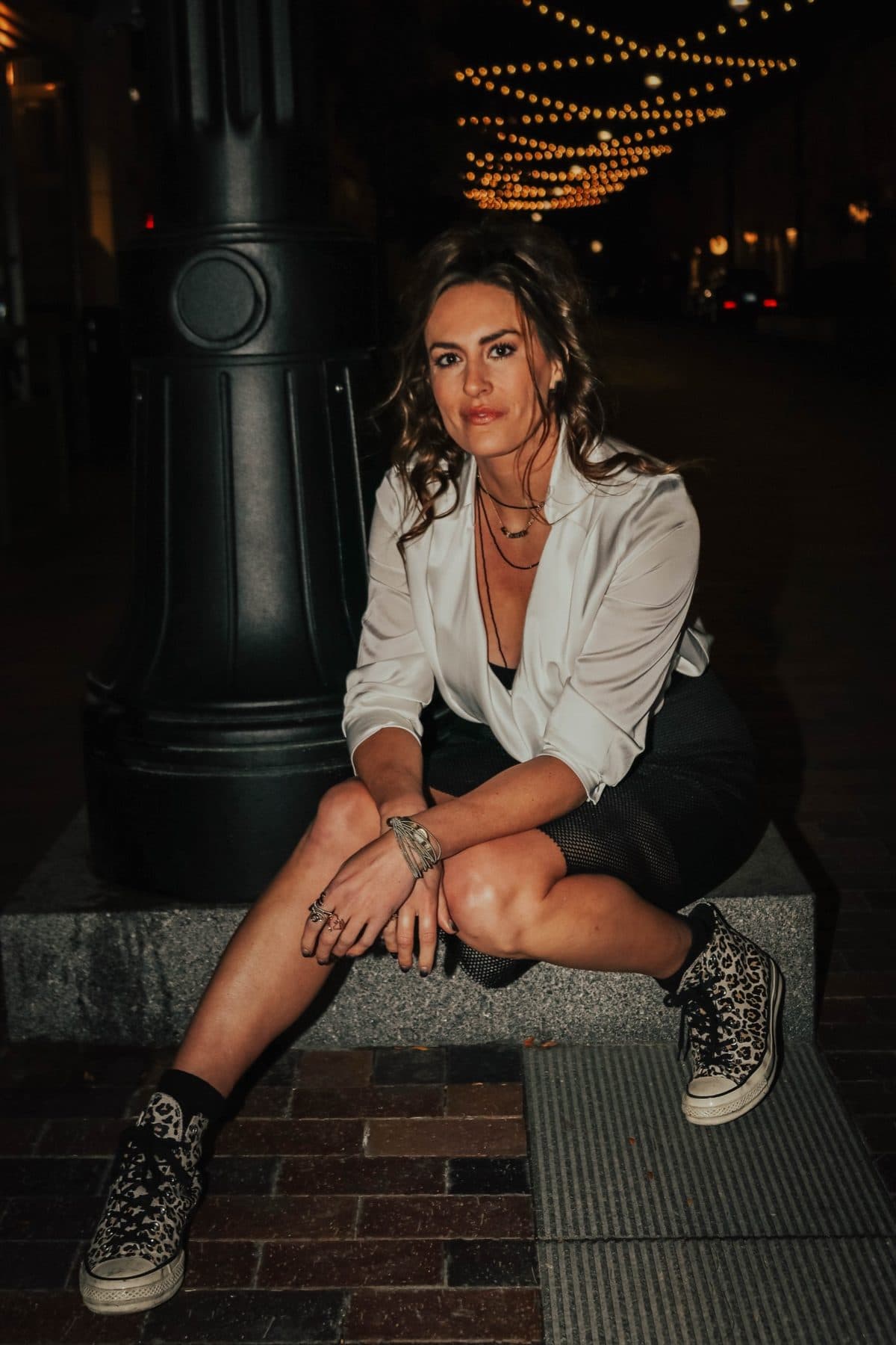 Crouched under string lights at night, white blazer and studded sneakers.