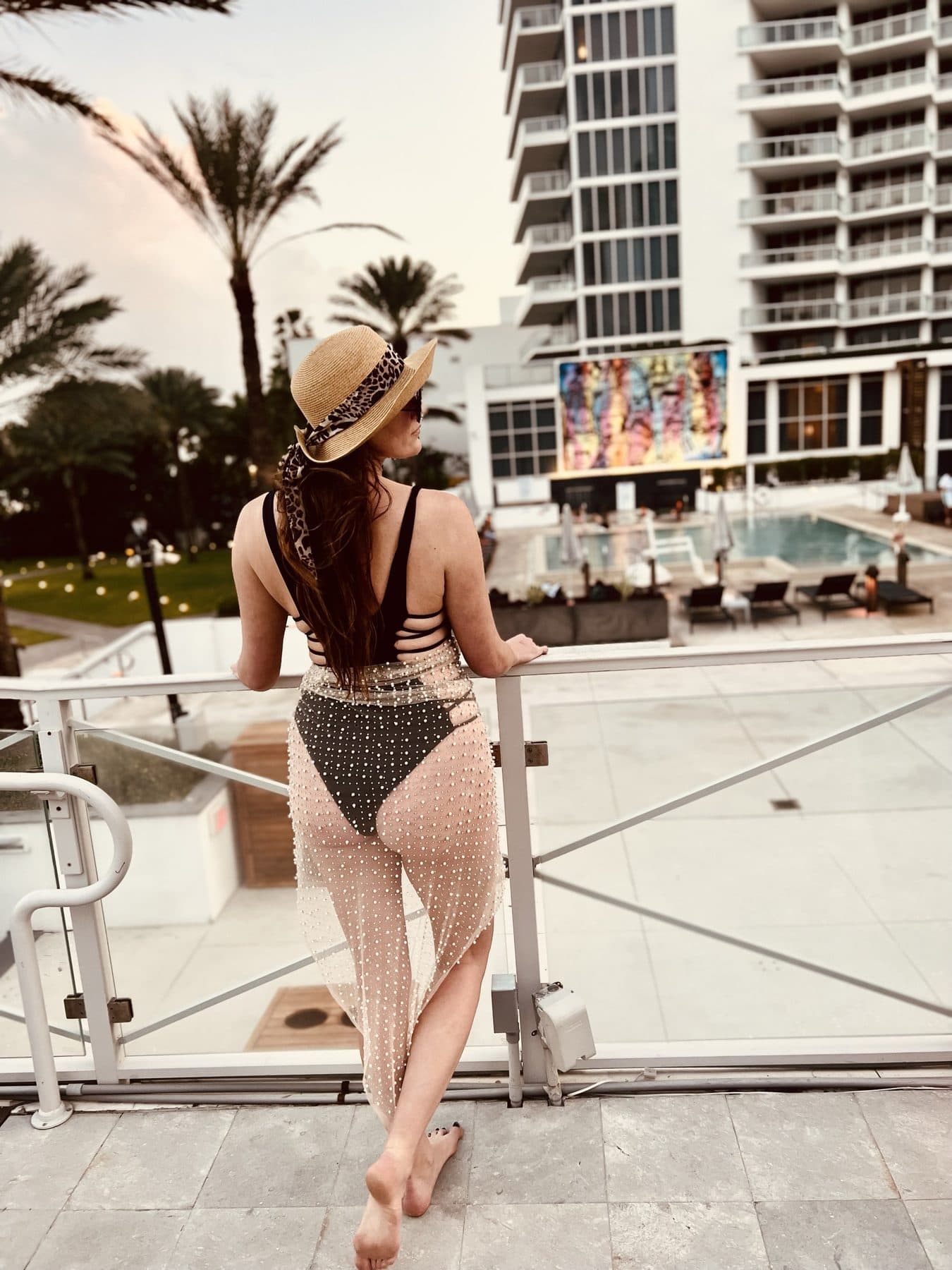 Back to the camera in a black one-piece and crochet wrap, palms and pool ahead.