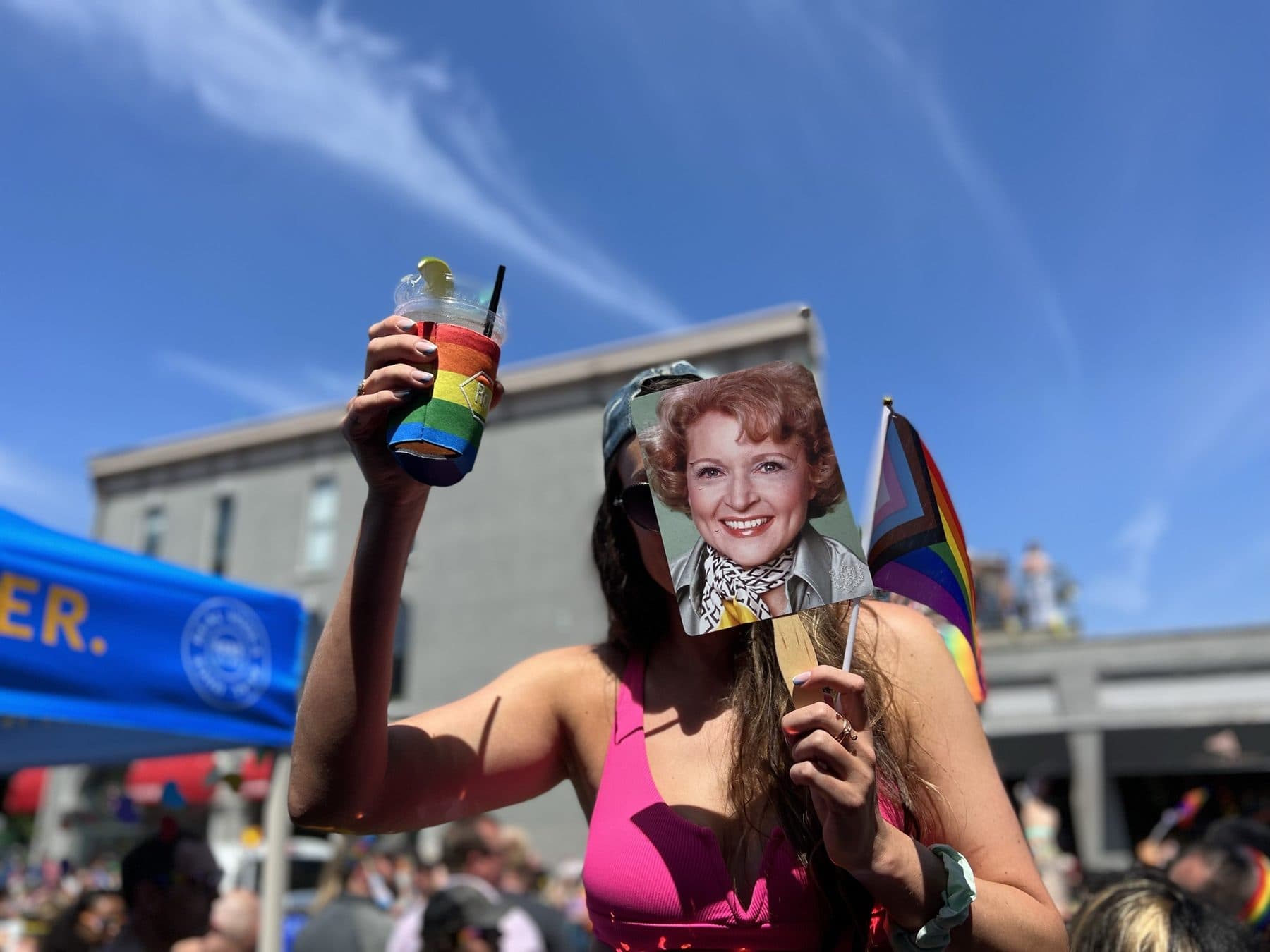 Pink top, rainbow drink raised, sunglasses on at a street festival.