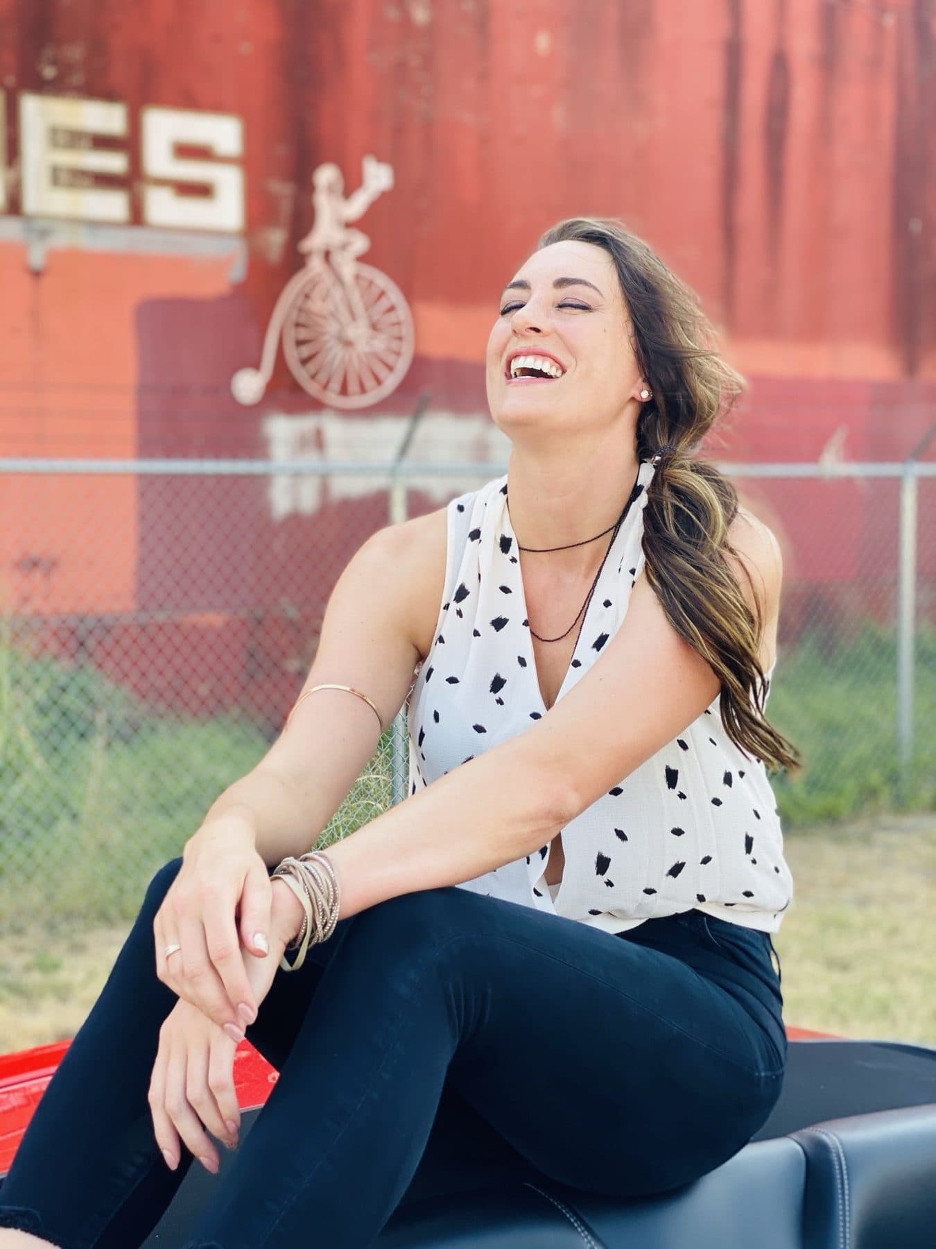 Laughing on the hood of a car in a dalmatian-print top and dark denim.