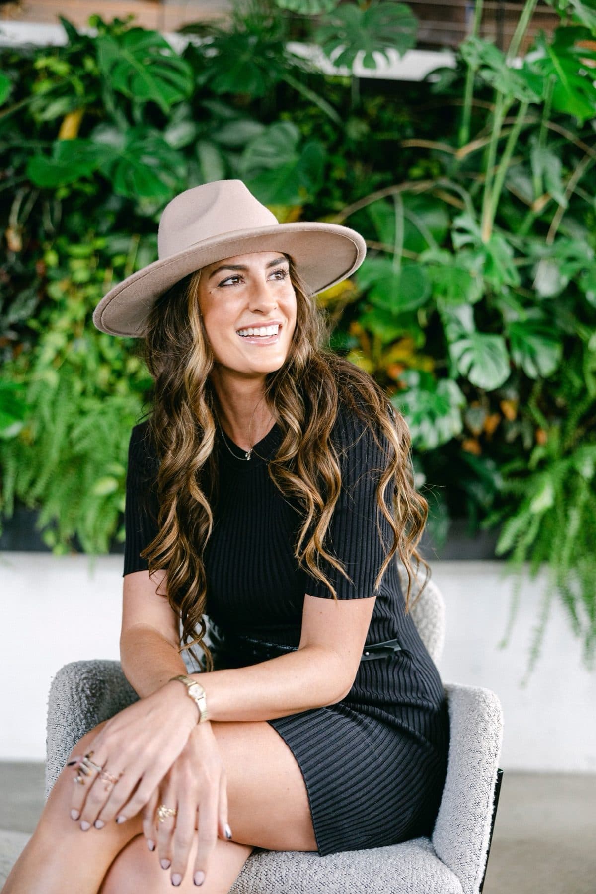 Seated in a black dress and cream hat against a living green wall.