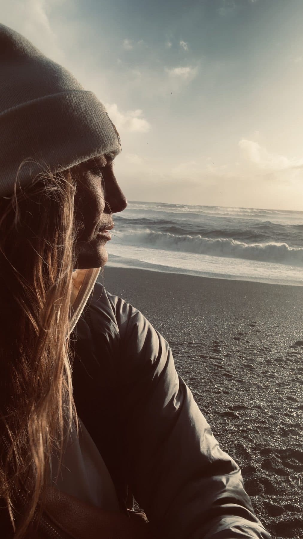 Beanie and jacket on a black-sand beach, profile to the surf.