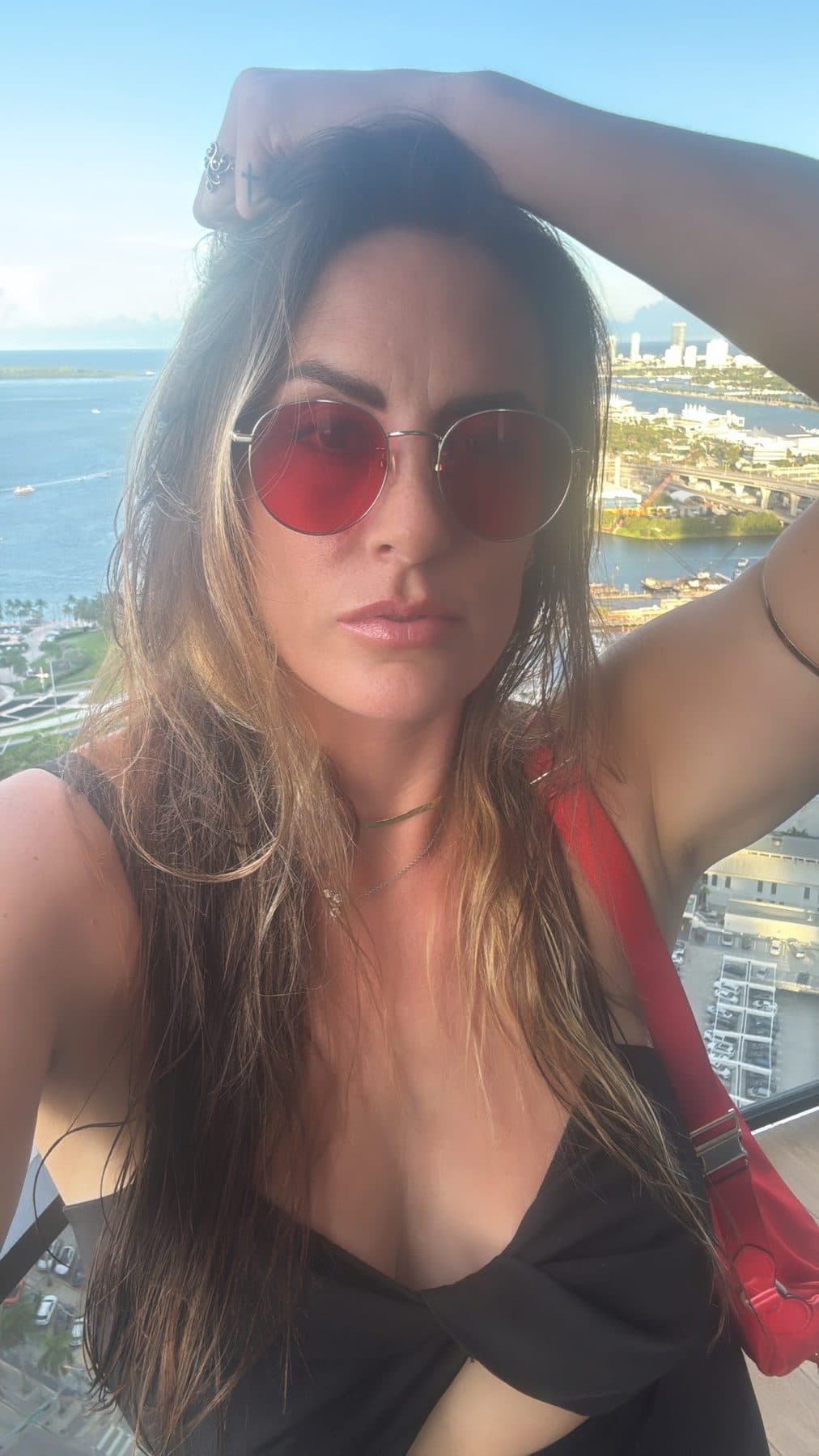 Rose-tinted sunglasses, hand in hair, sea and skyline behind.