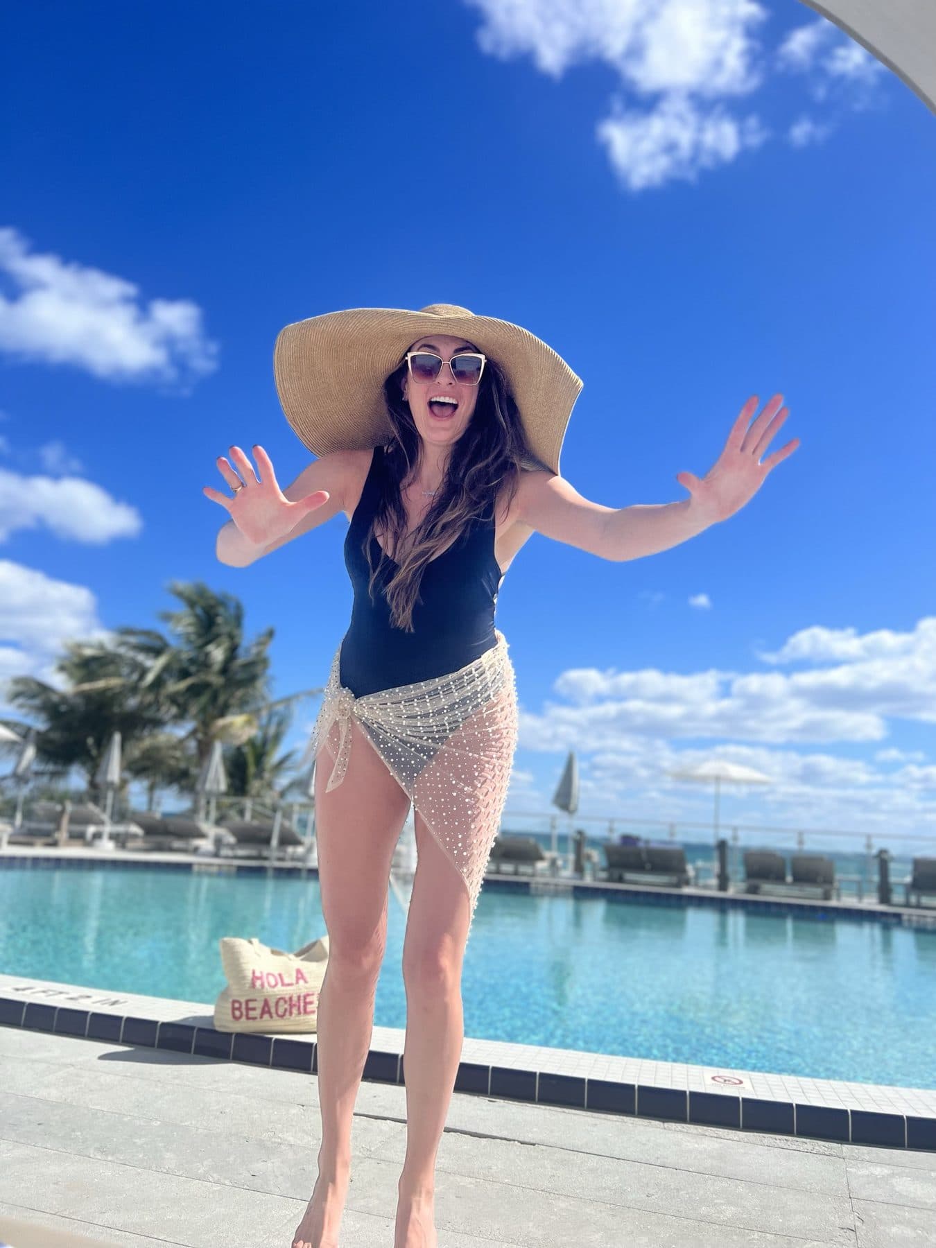 Wide straw hat, arms up, poolside under a blue sky.