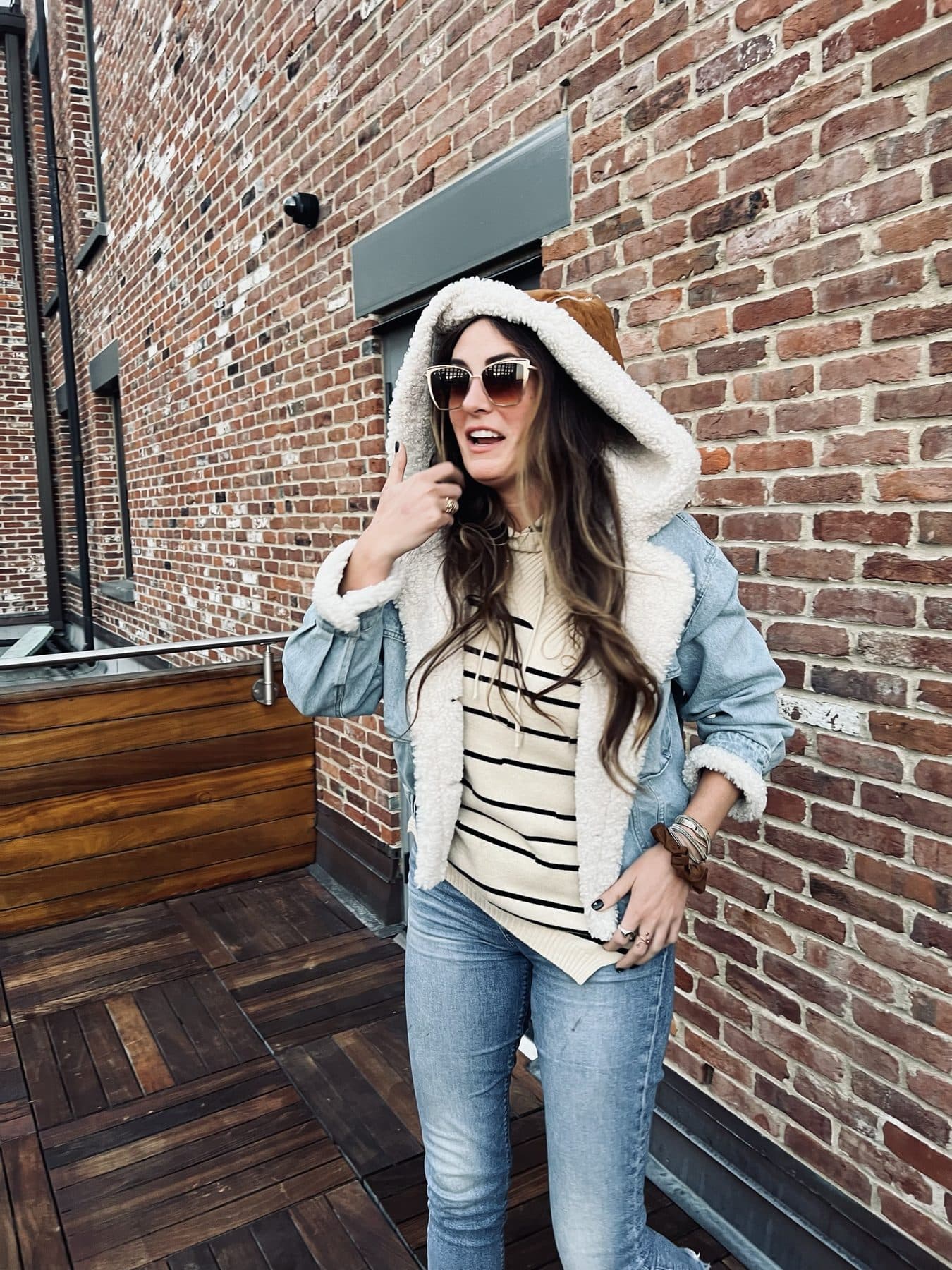 Shearling-lined denim jacket, hood up, against a red brick wall.