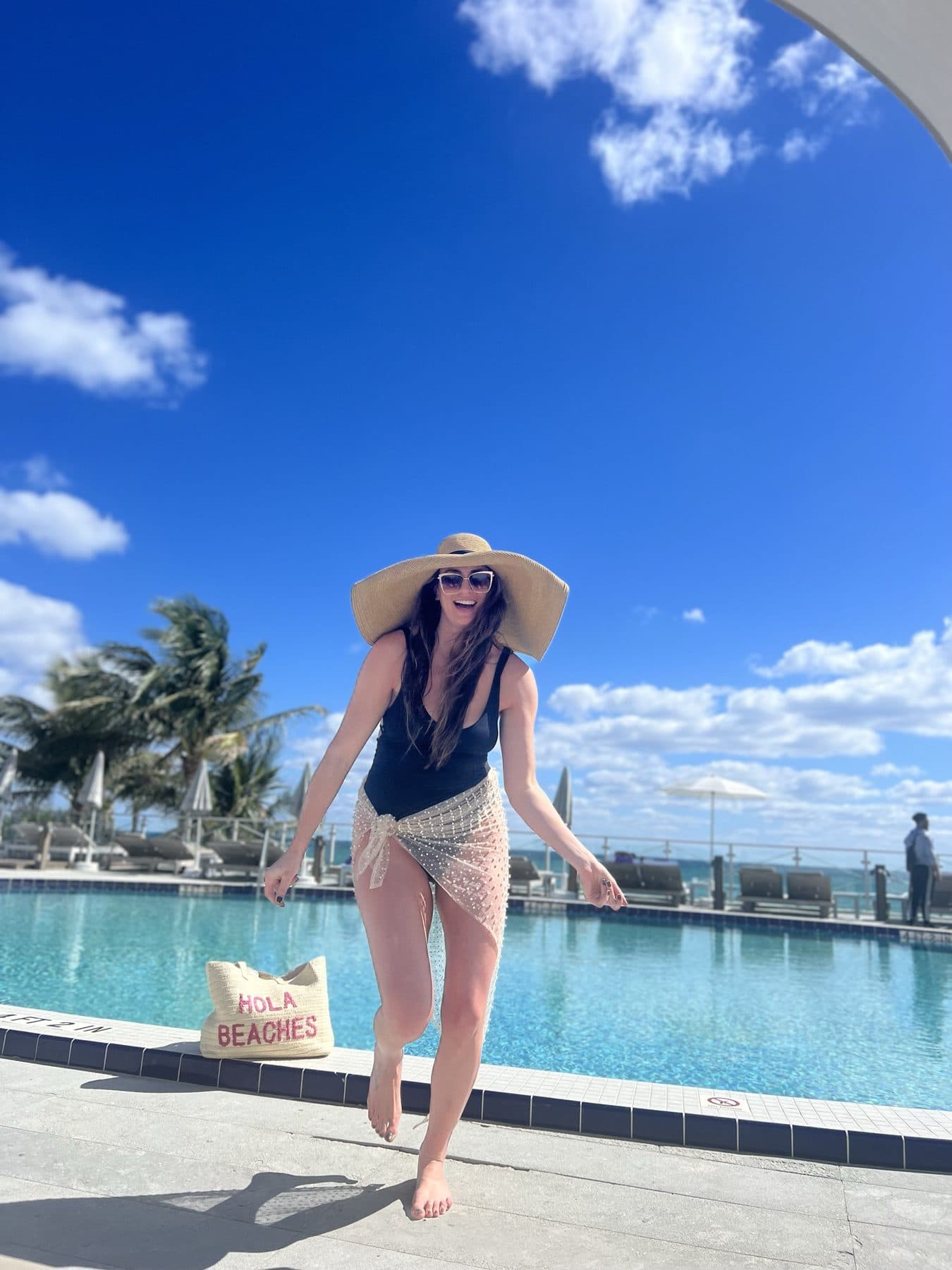 Barefoot beside the pool in a black one-piece and straw hat.
