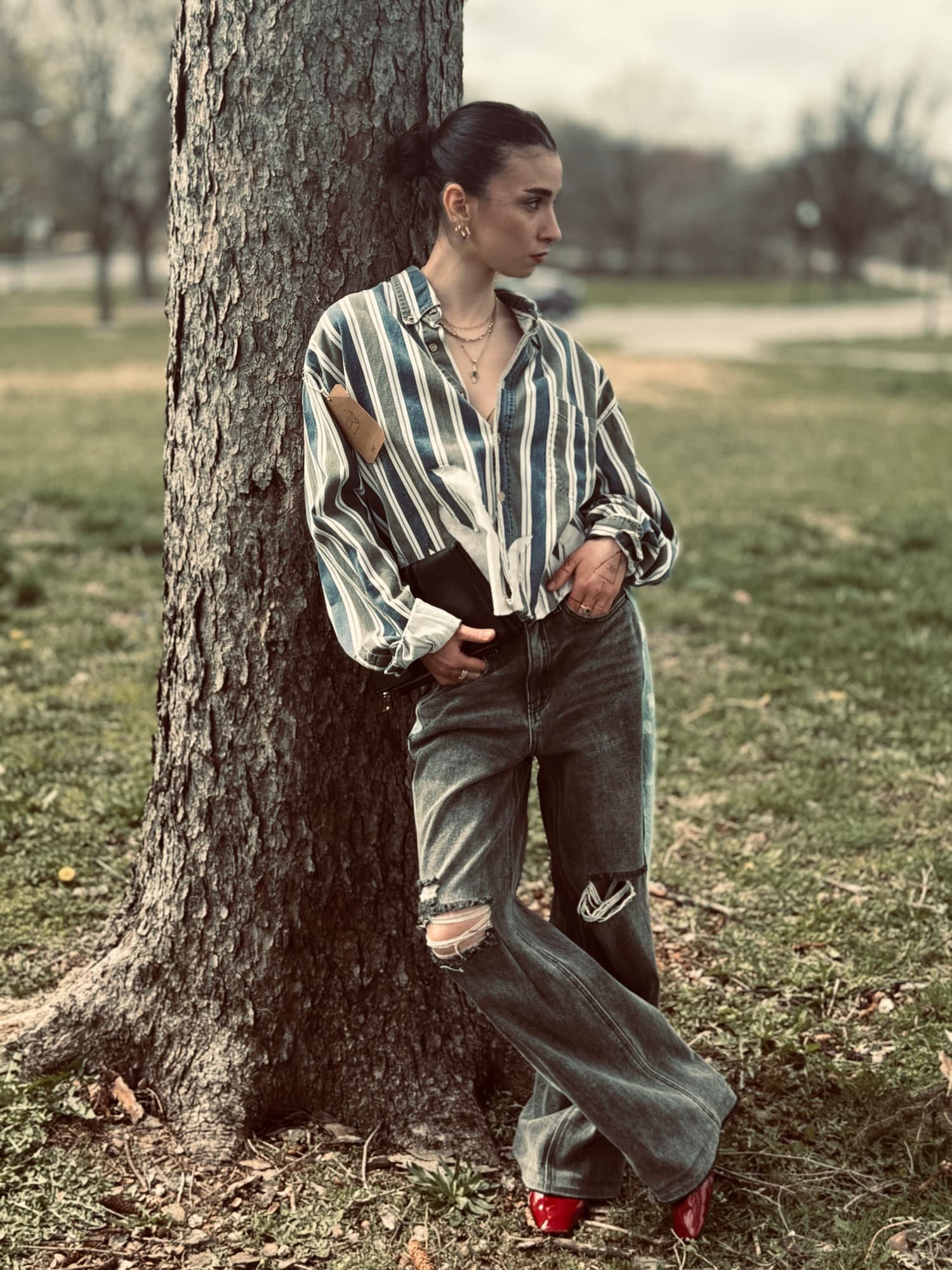 Haley leaning against a tree trunk in a park, oversized striped button-down, wide-leg distressed jeans, and glossy red shoes, gaze off to the side.