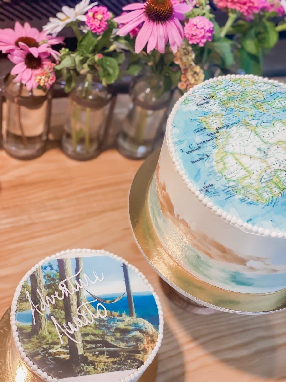 The watercolor world-map cake beside a smaller hammock-scene cake, with pink coneflowers in amber bud vases.