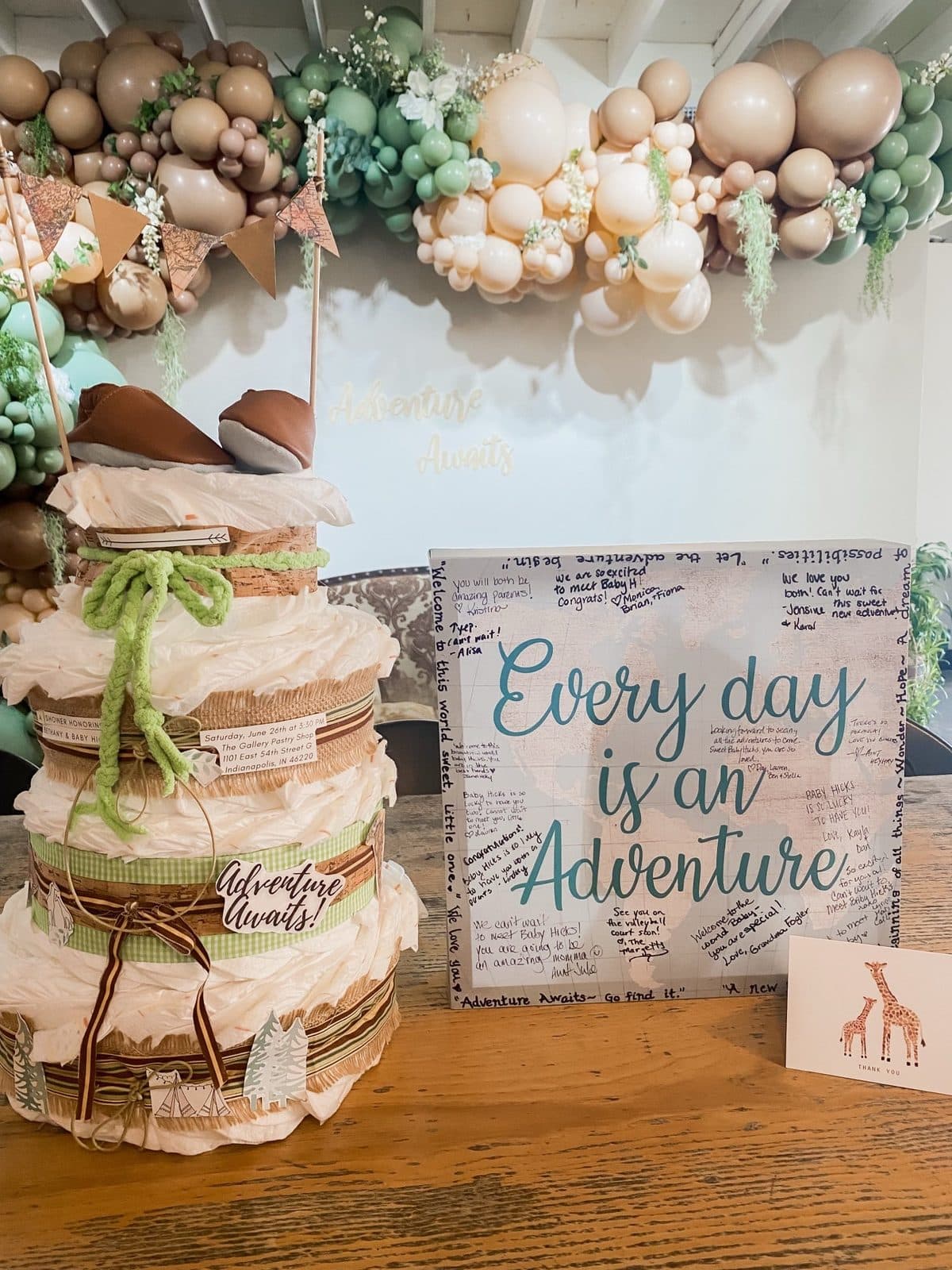 A diaper cake with a sage ribbon beside an 'Every day is an Adventure' guest-signing board, under the balloon garland.