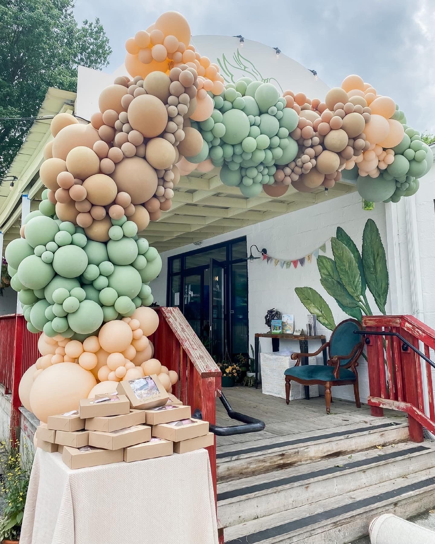 The venue entrance wrapped in a sage-and-clay balloon arch over a favor-box display, set against a painted botanical mural.