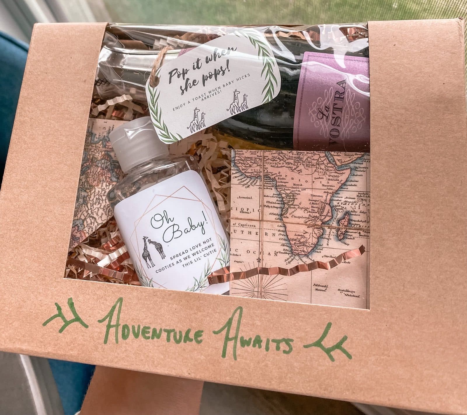 A kraft favor box stamped 'Adventure Awaits' holding a mini 'Oh Baby' hand sanitizer, a map-print card, and a treat.