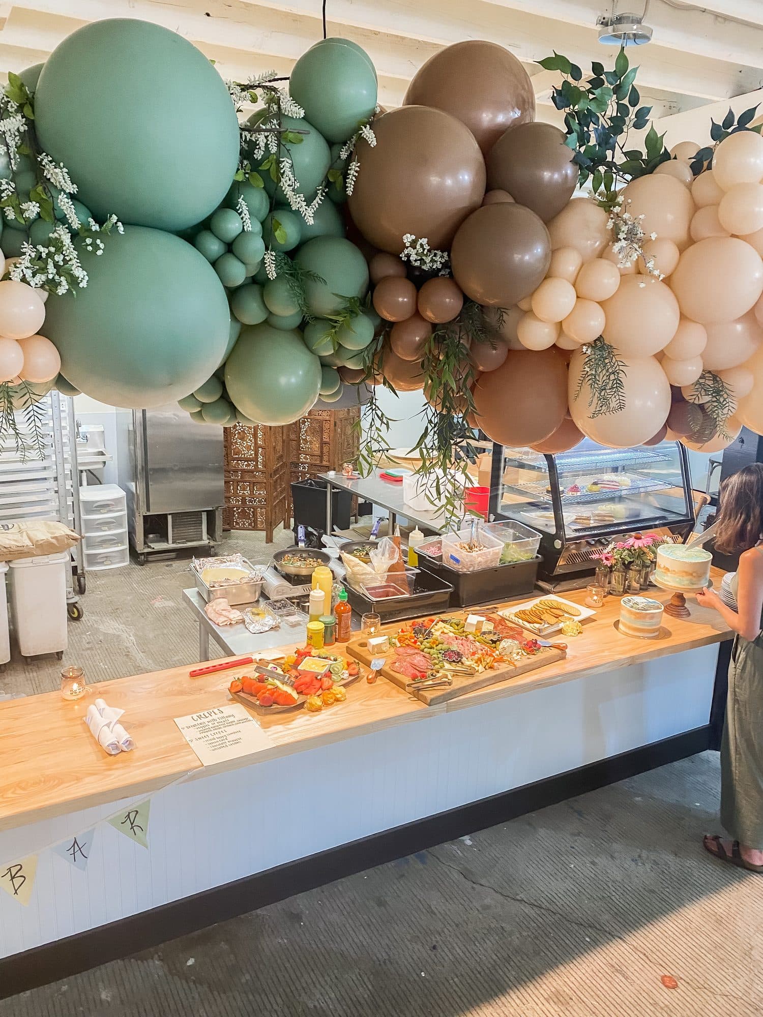 A grazing table of charcuterie and fruit boards under a sage-and-brown balloon installation, with a naked cake at the end.