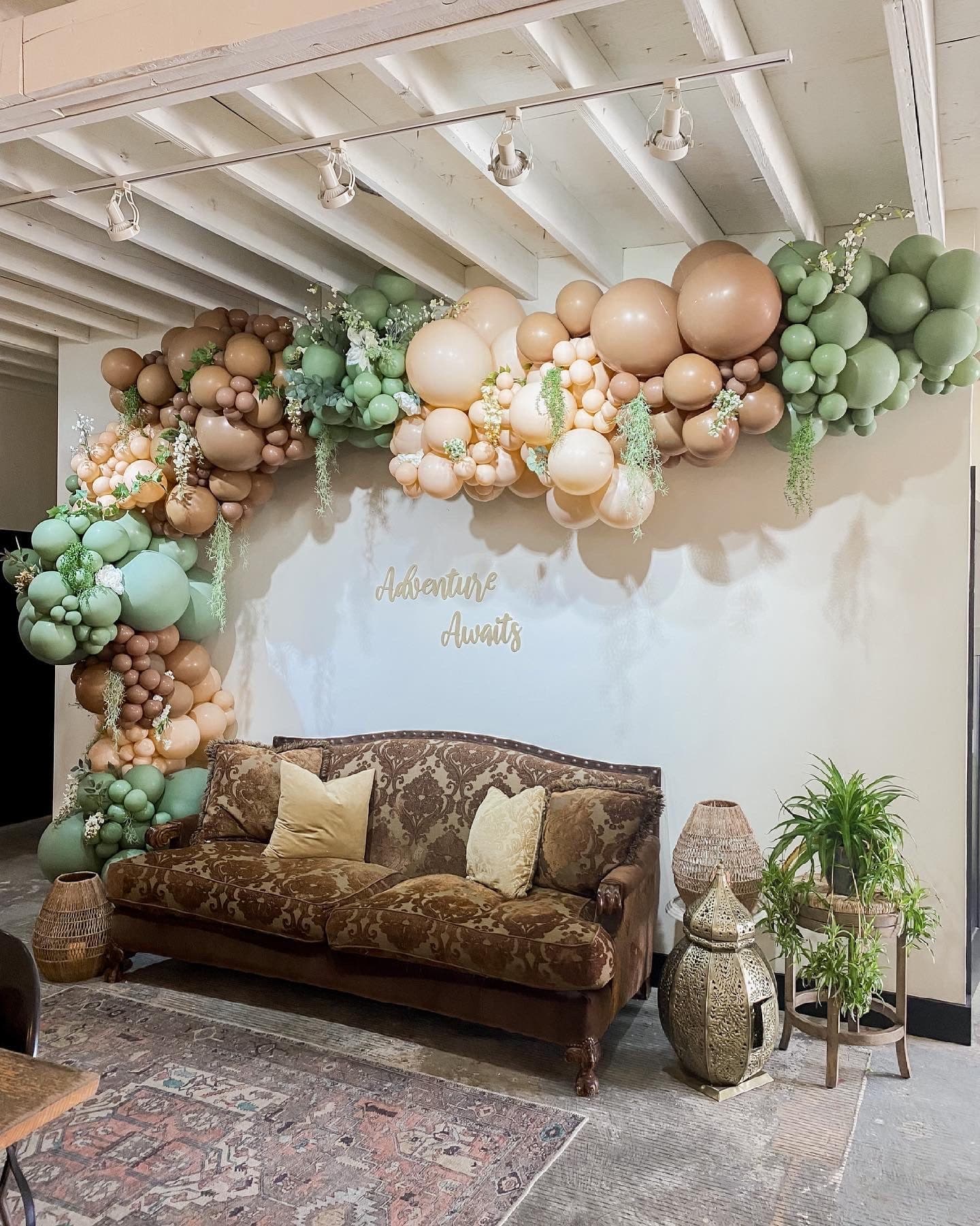A sage, cream, and tan balloon arch over a vintage brown velvet sofa with an Adventure Awaits script sign.