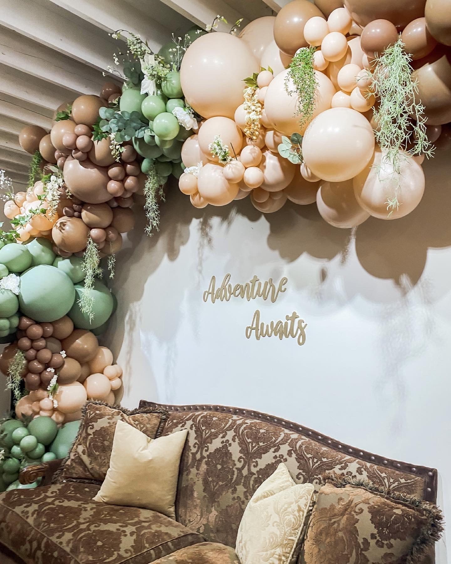 A sage, sand, and clay balloon arc with trailing greenery and dried botanicals framing a damask settee and gold 'Adventure Awaits' lettering.