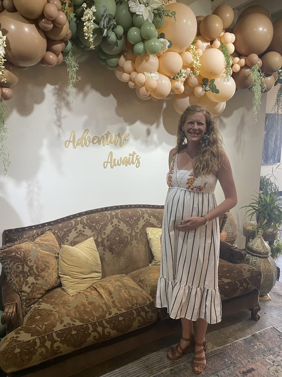 The mom-to-be in a striped sundress beside the damask settee, one hand on her bump, under the balloon garland and 'Adventure Awaits' script.