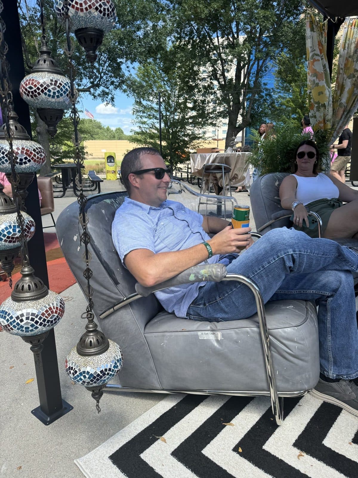 A guest kicked back in a grey leather club chair beside a mosaic lantern, umbrellas and trees behind.