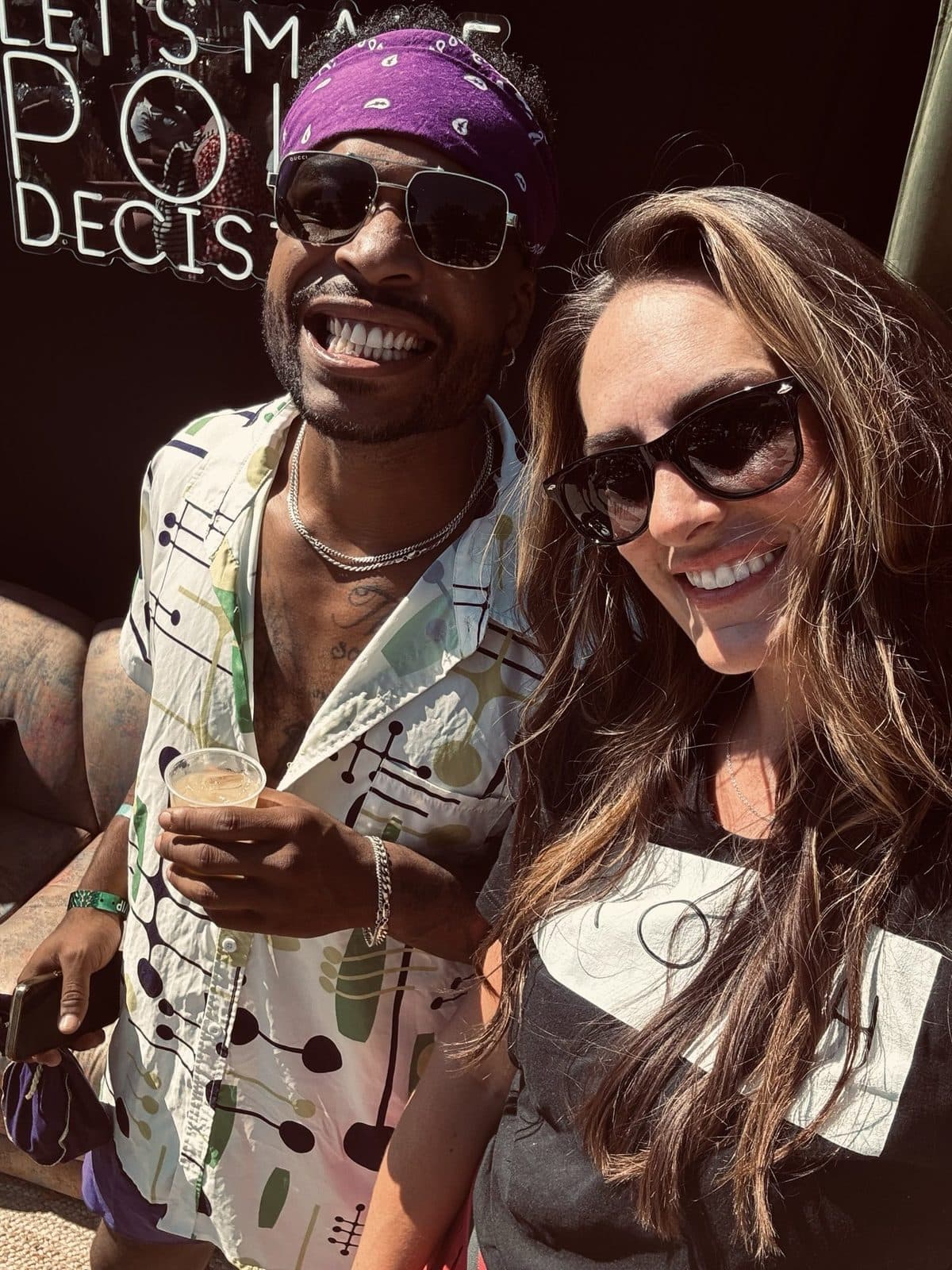 Two friends in sunglasses grinning under the neon sign, a purple bandana and a name-tag sticker.