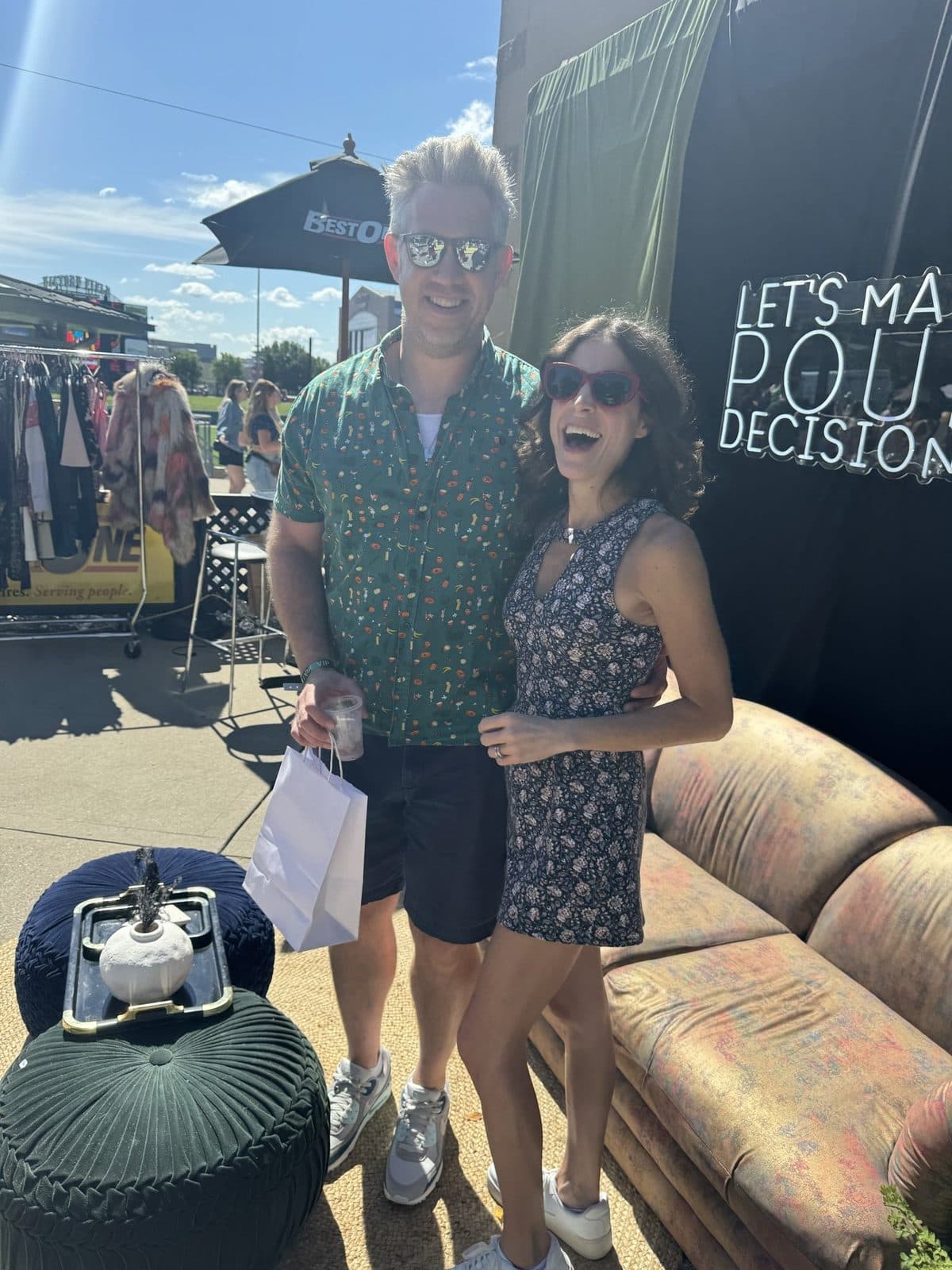 A couple in matching floral prints laughing in front of the 'Let's Make Pour Decisions' backdrop, the vintage sofa beside them.