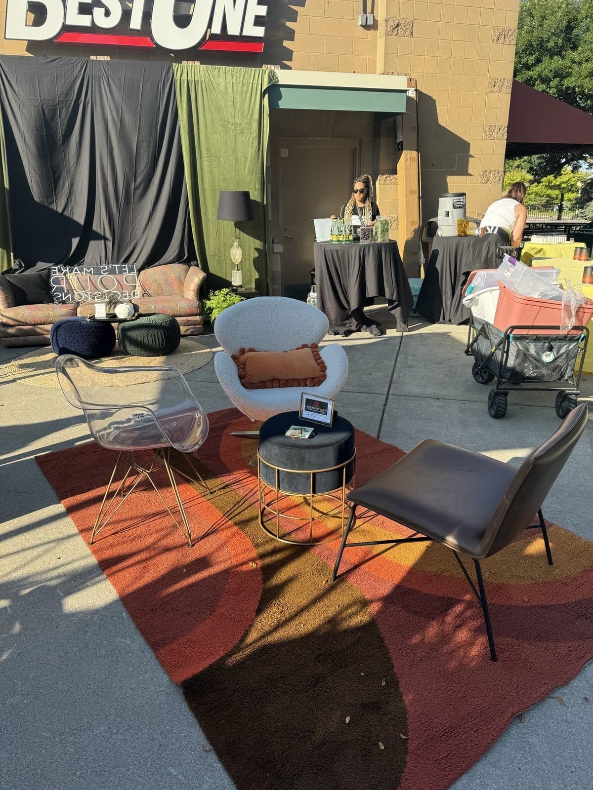 The lounge staged on a painted concrete lot, ghost chairs and a clay accent pillow around a gold side table, beneath a BestOne building sign.