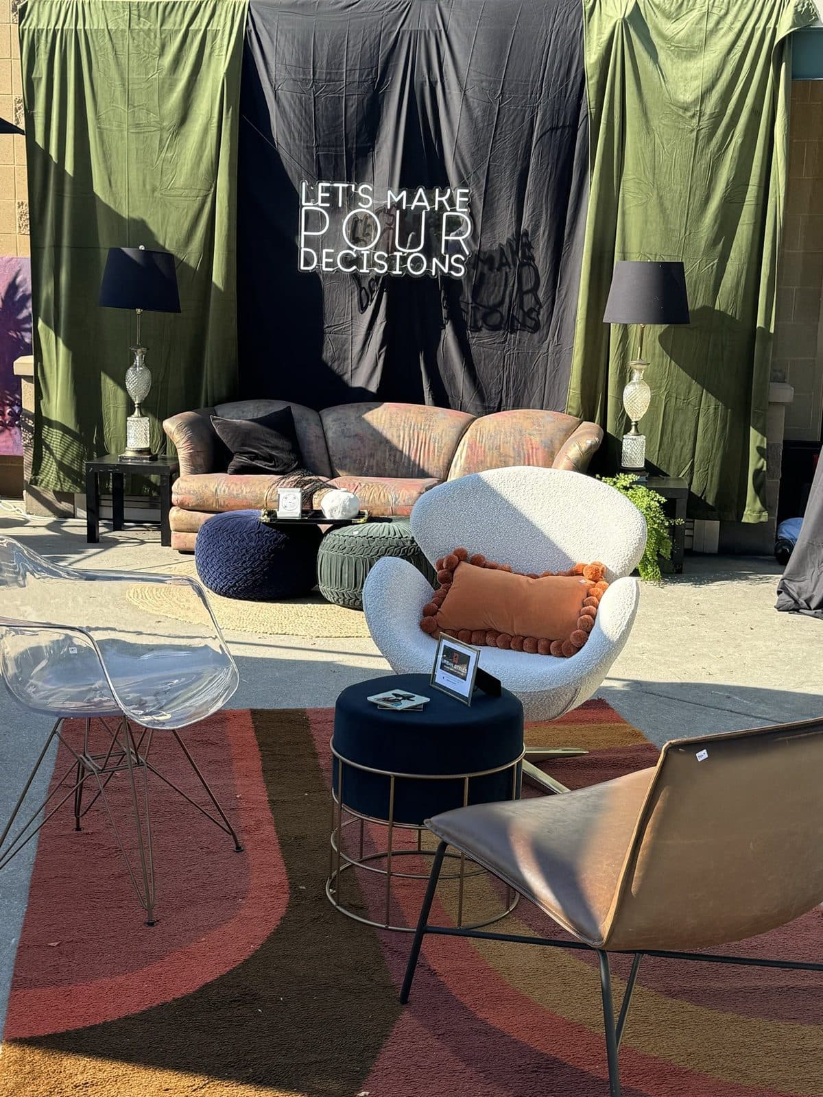 The VIP lounge under a 'Let's Make Pour Decisions' sign: a white egg chair, clear ghost chairs, a sage bouclé pouf, and a velvet sofa on a rust-and-cream painted lot, draped in olive and black.