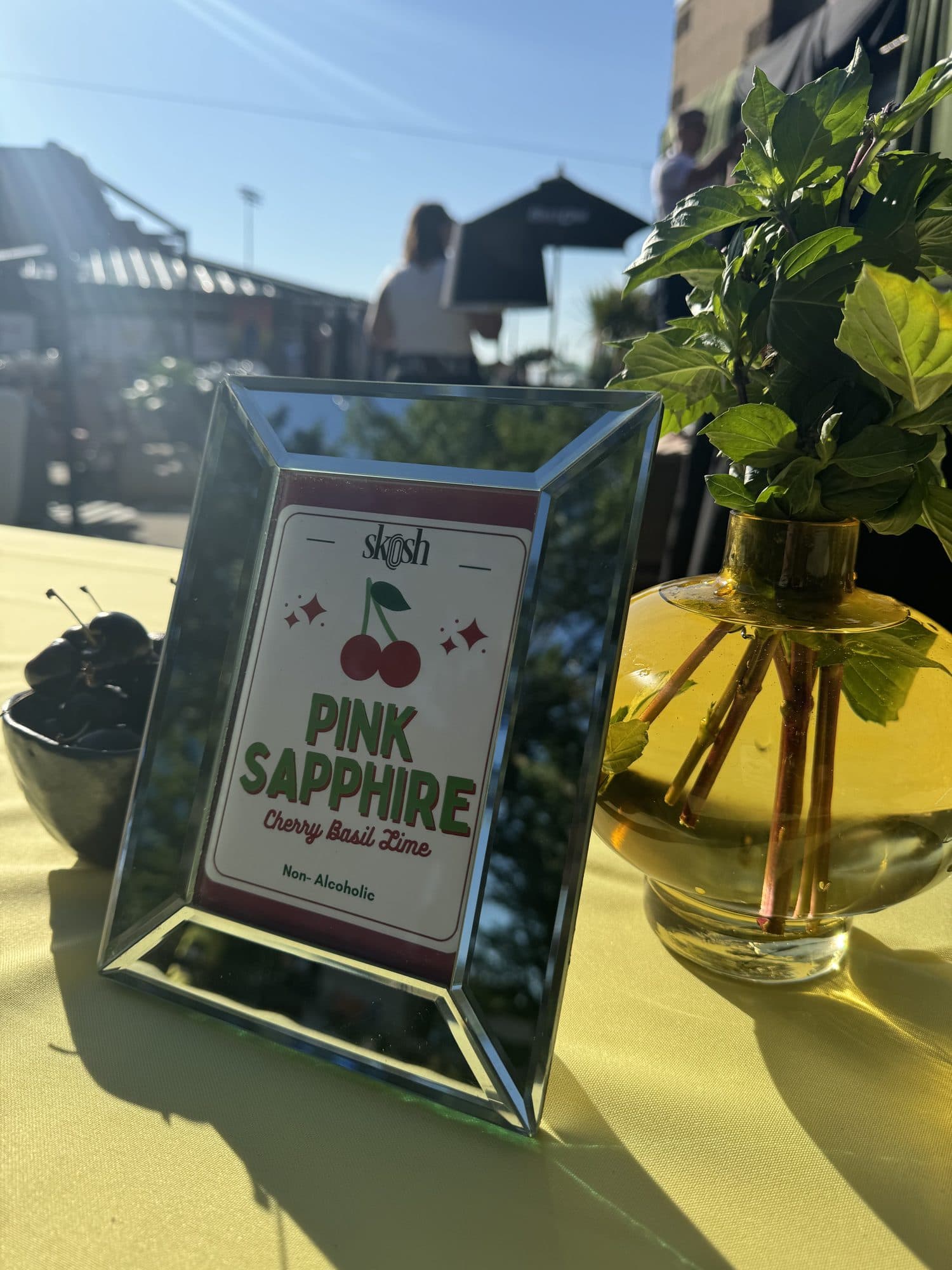 A mirrored frame showing Skosh's 'Pink Sapphire, Cherry Basil Lime, Non-Alcoholic' beside a gold vase of fresh mint on a butter-yellow cloth.