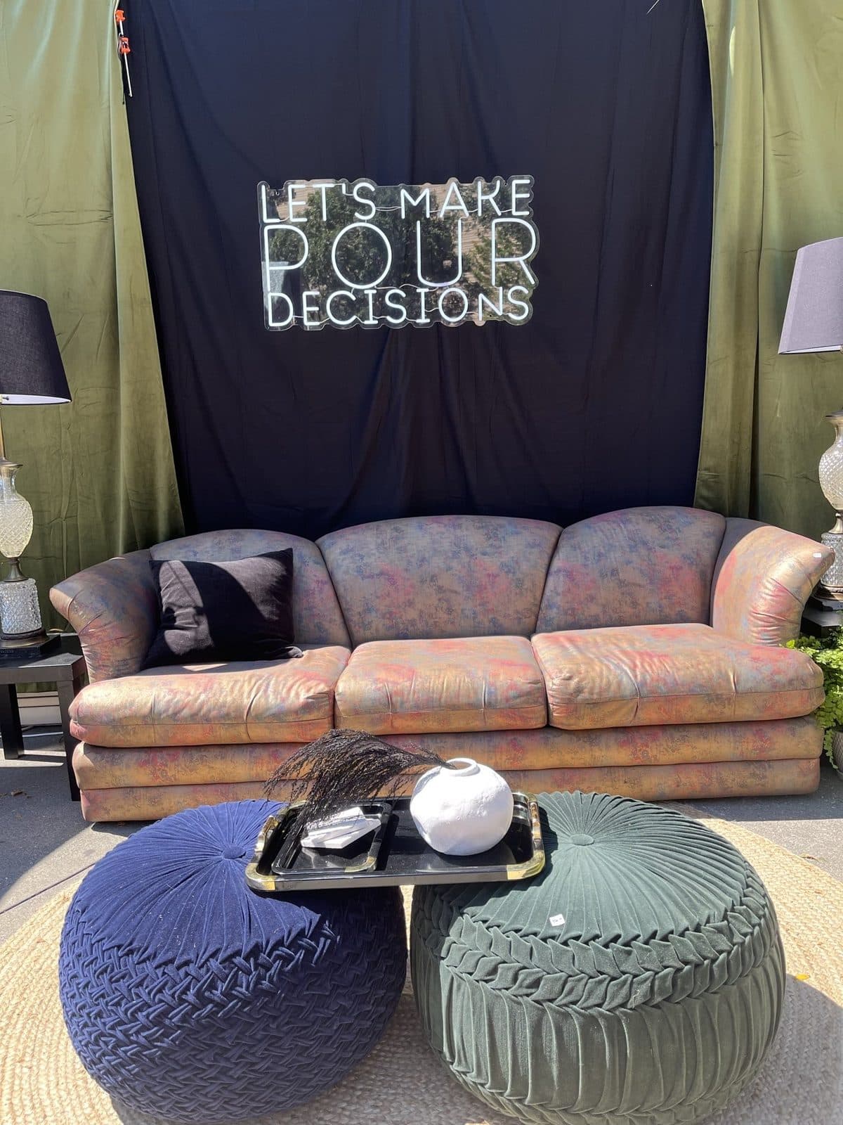 The vintage floral velvet sofa with a navy and a sage bouclé pouf on a layered rug under the neon sign.
