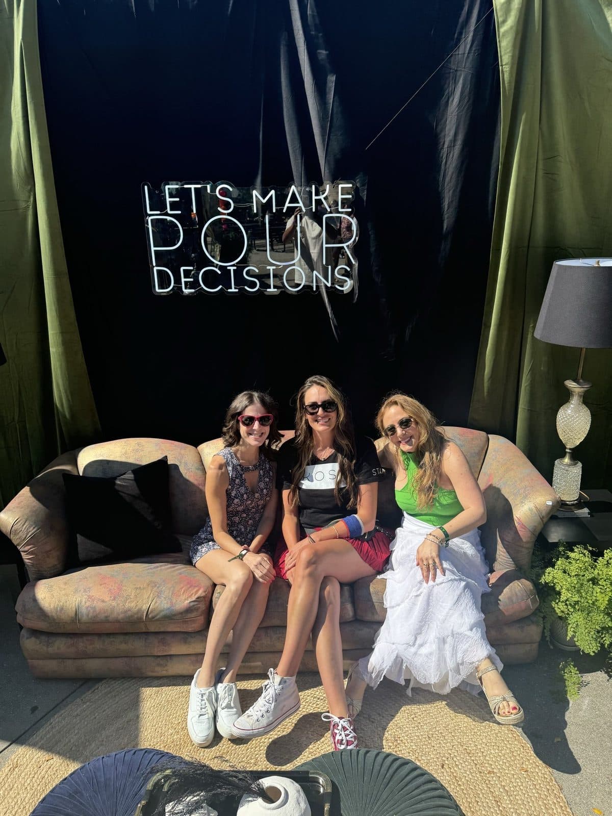 Three guests on the vintage velvet sofa under the neon 'Let's Make Pour Decisions' sign, drinks in hand.