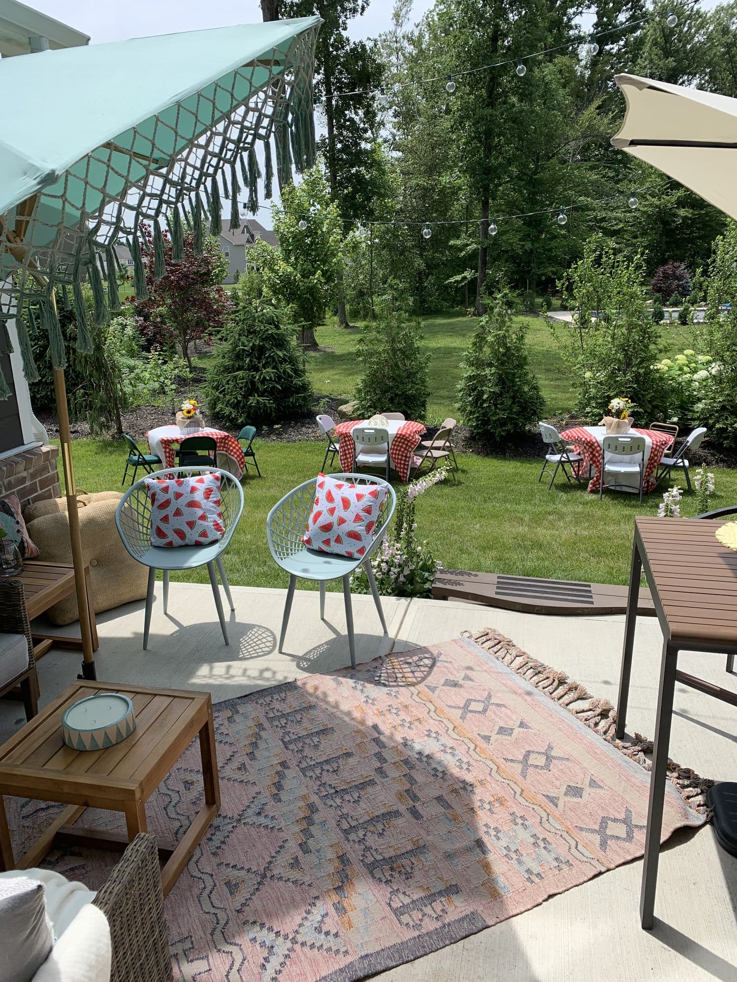 Backyard tables dressed in red gingham with watermelon-print pillows on rattan chairs, umbrellas, and a layered outdoor rug.