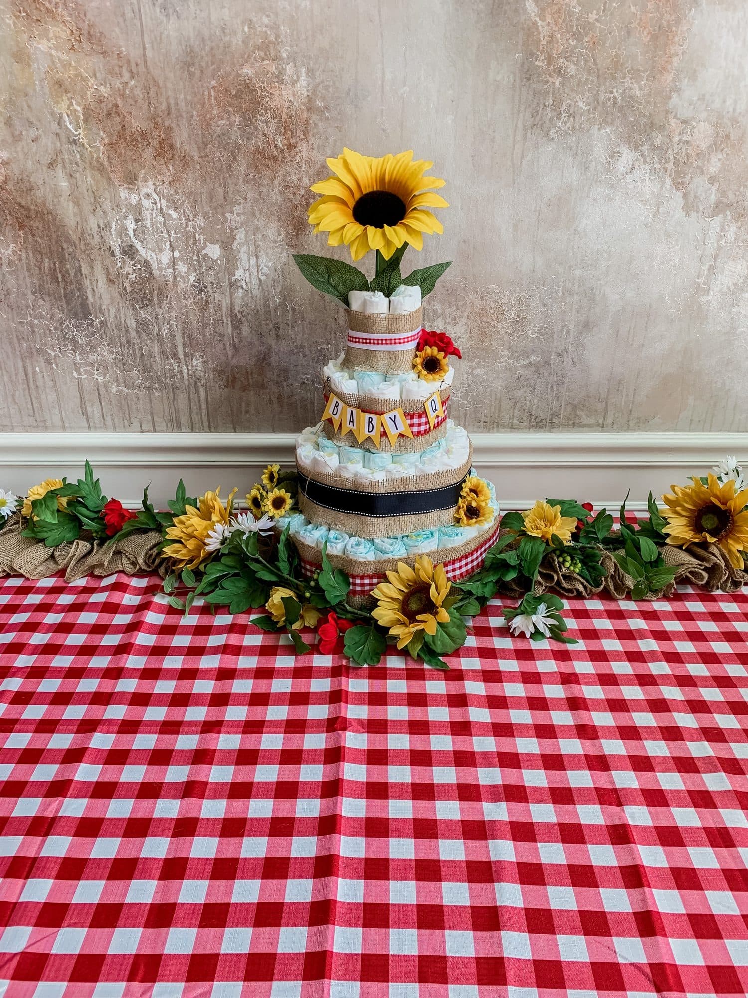 A burlap-and-gingham diaper cake topped with a sunflower, set on a red-gingham cloth against a textured backdrop.