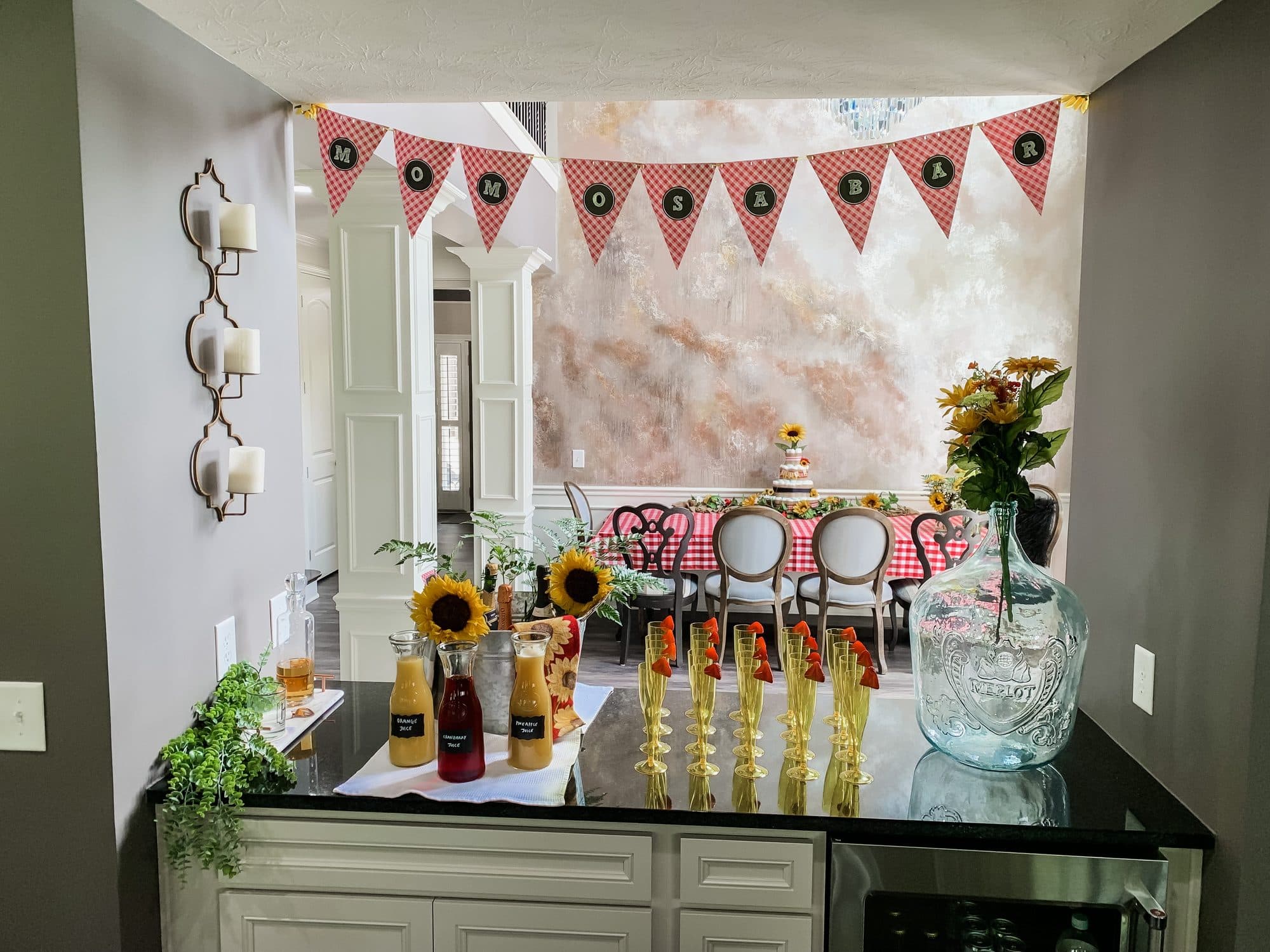 A 'Momosa Bar' with red-gingham pennant bunting, juices in glass carafes, sunflowers, and amber flutes, with a gingham-set dining table beyond.