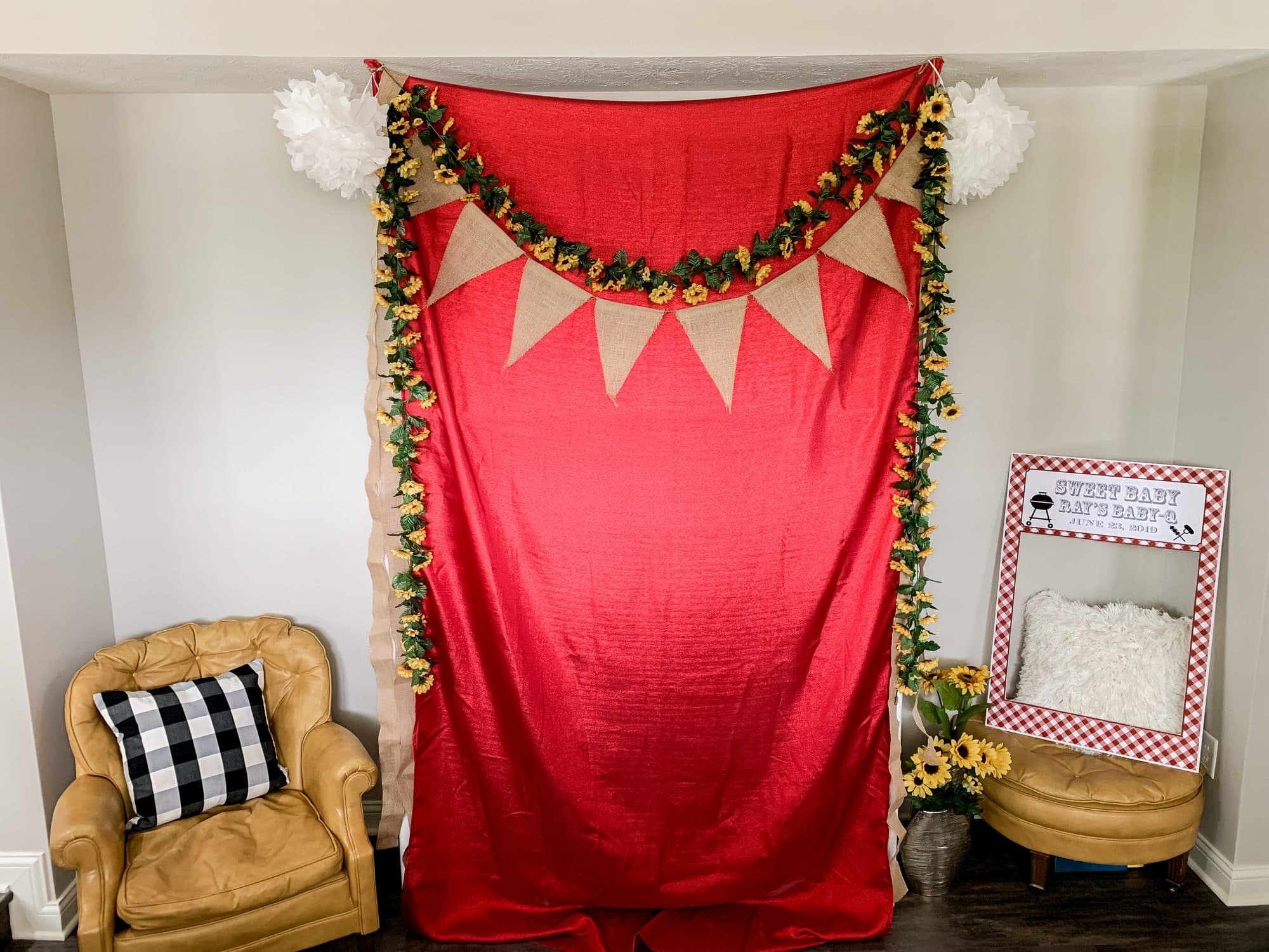 A photo-booth corner, a red backdrop with sunflower-and-burlap garland, a gingham selfie frame, and tan leather seating.