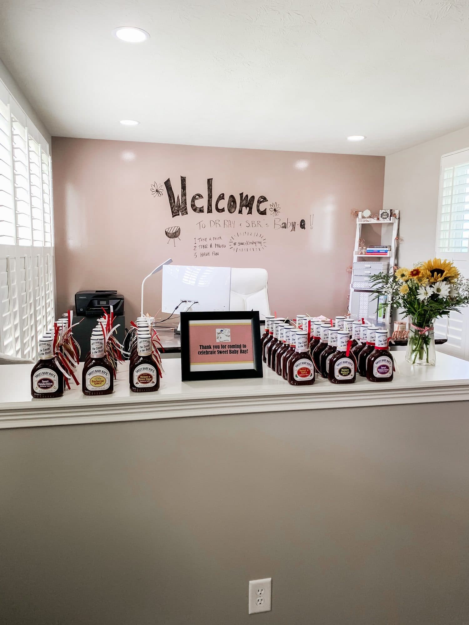A welcome table of Sweet Baby Ray's barbecue-sauce bottle favors with ribbon tags, a framed thank-you sign, and a sunflower bouquet.