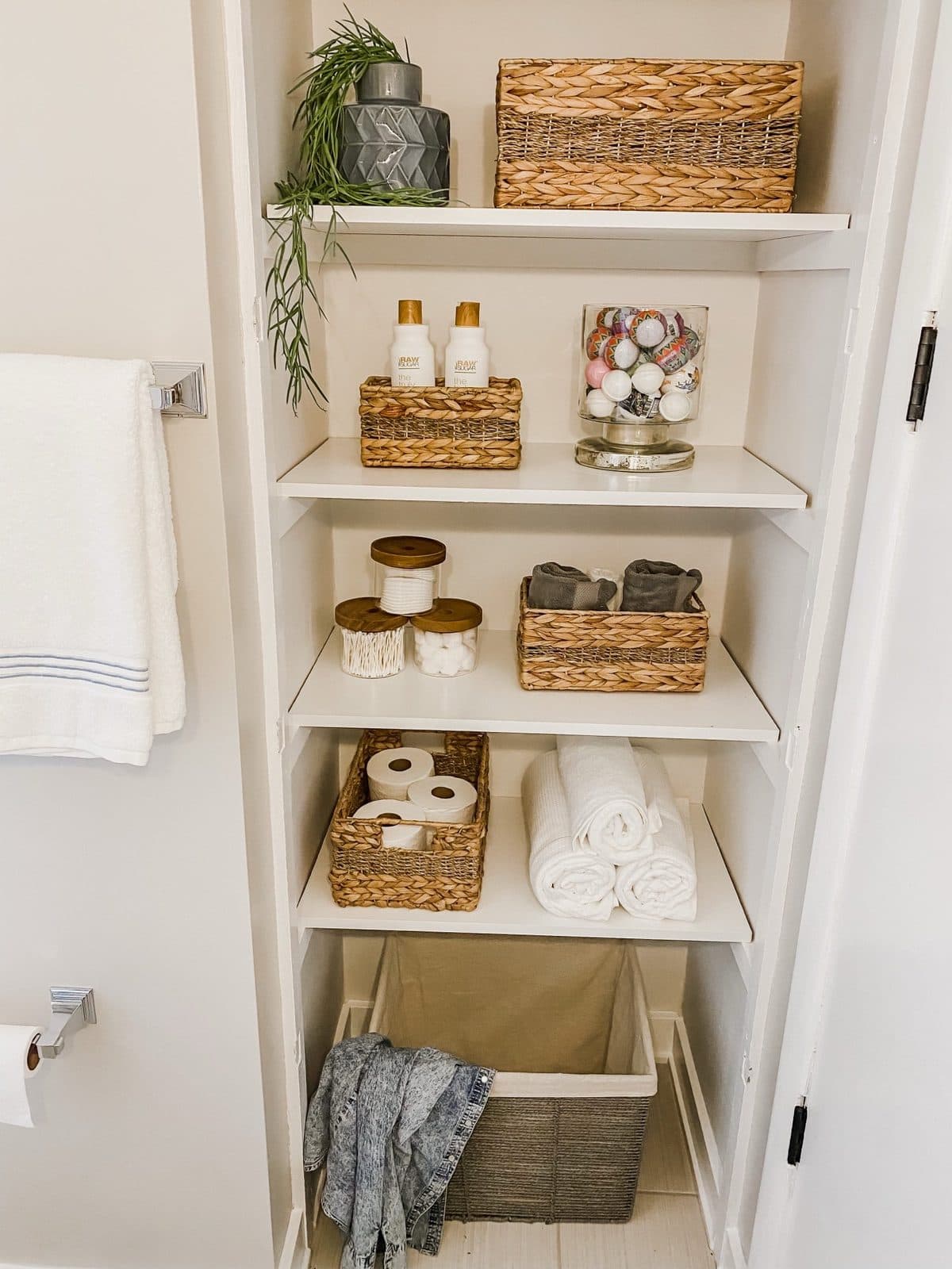 A linen closet reset: woven baskets, folded towels, and glass jars lined up on white shelves behind an open door.