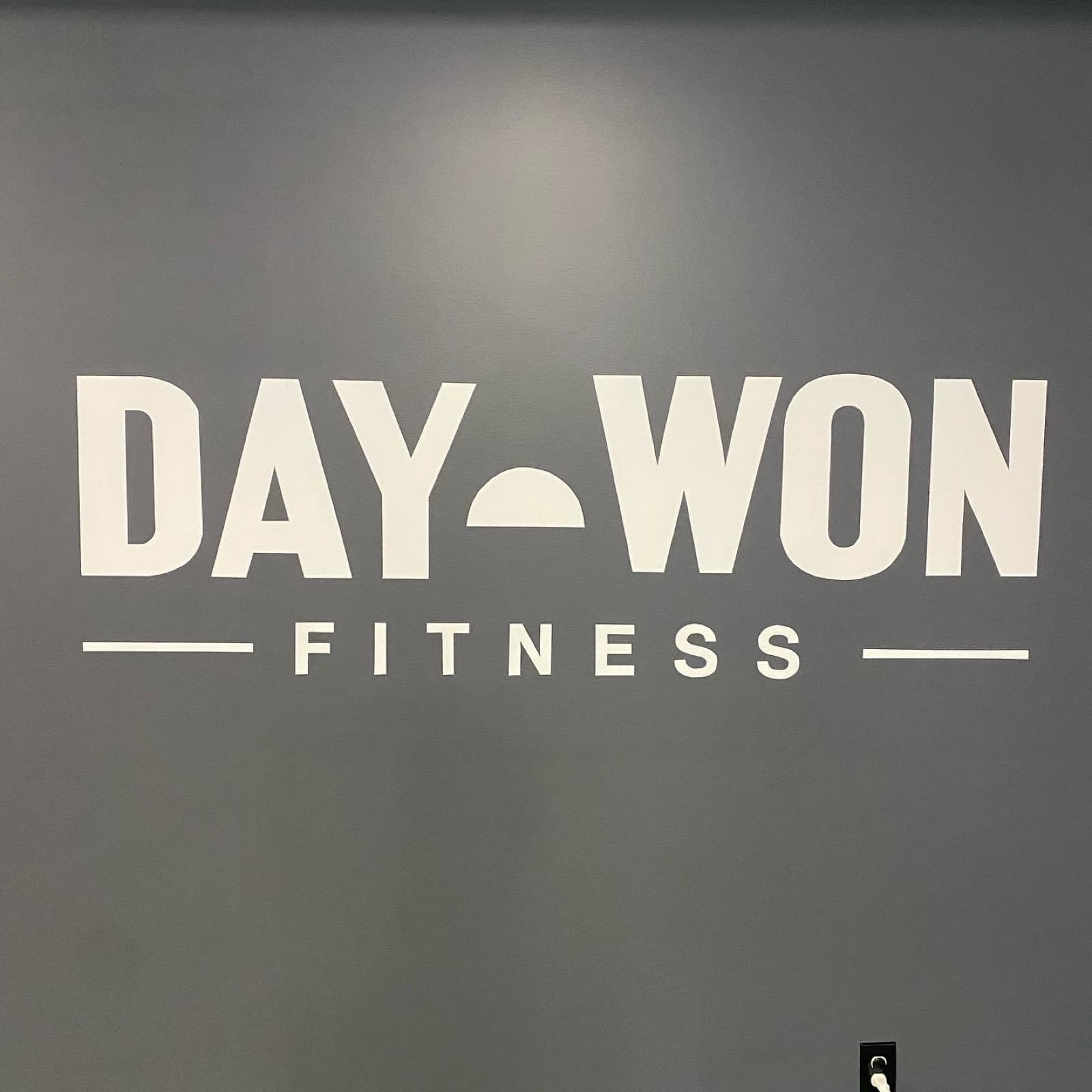 The hand-painted Day Won Fitness wordmark in crisp white on a slate-gray gym wall.