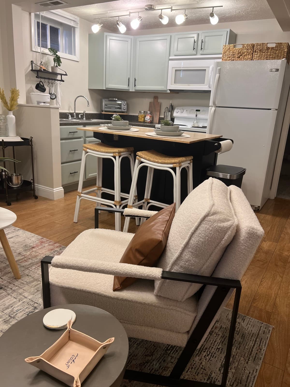 Sage-green galley kitchen with butcher-block island, rattan-and-white stools, and floating shelf styling.