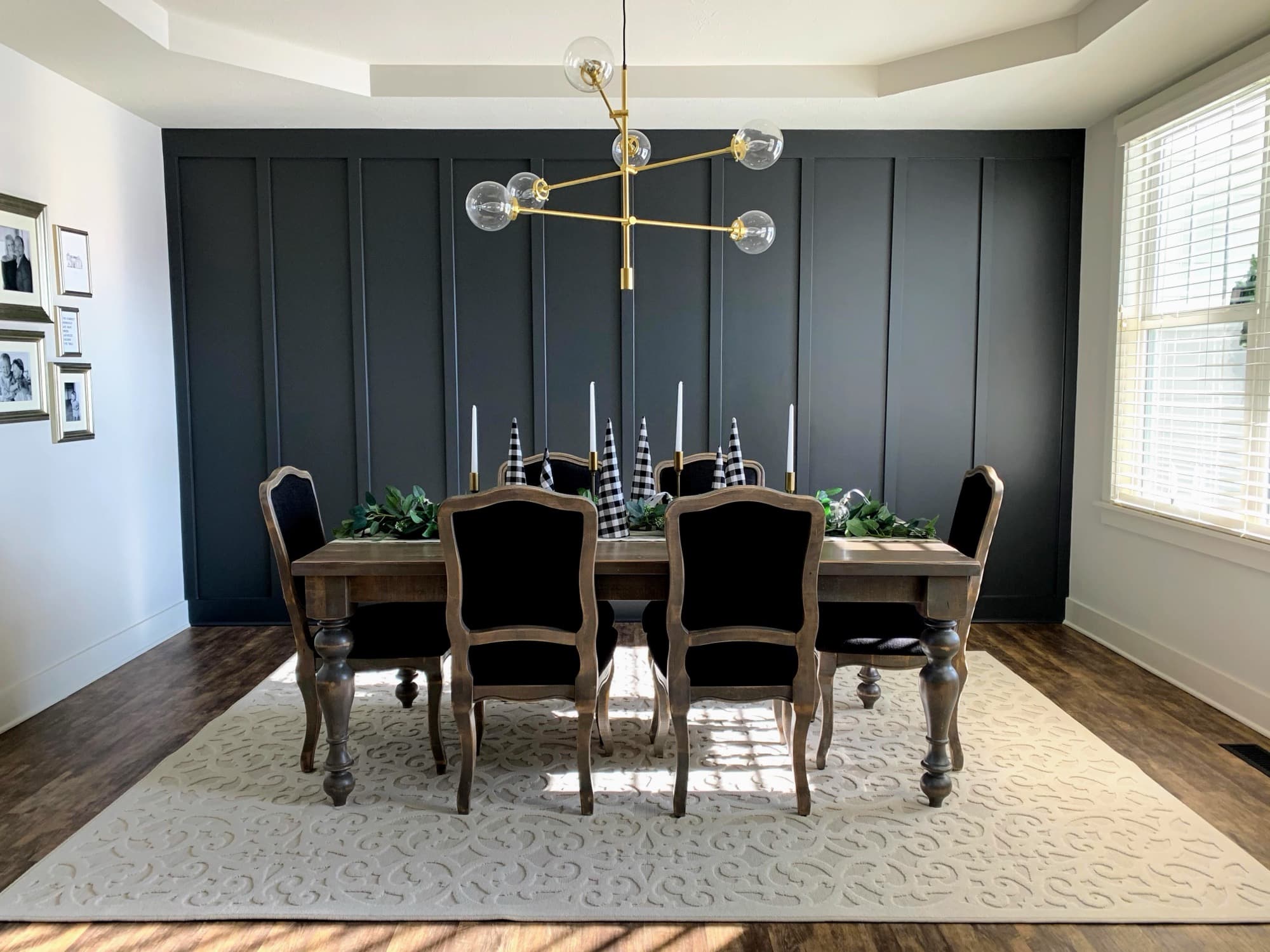 A floor-to-ceiling charcoal board-and-batten feature wall behind a walnut dining table, lit by directional window light, with a brass globe chandelier above.