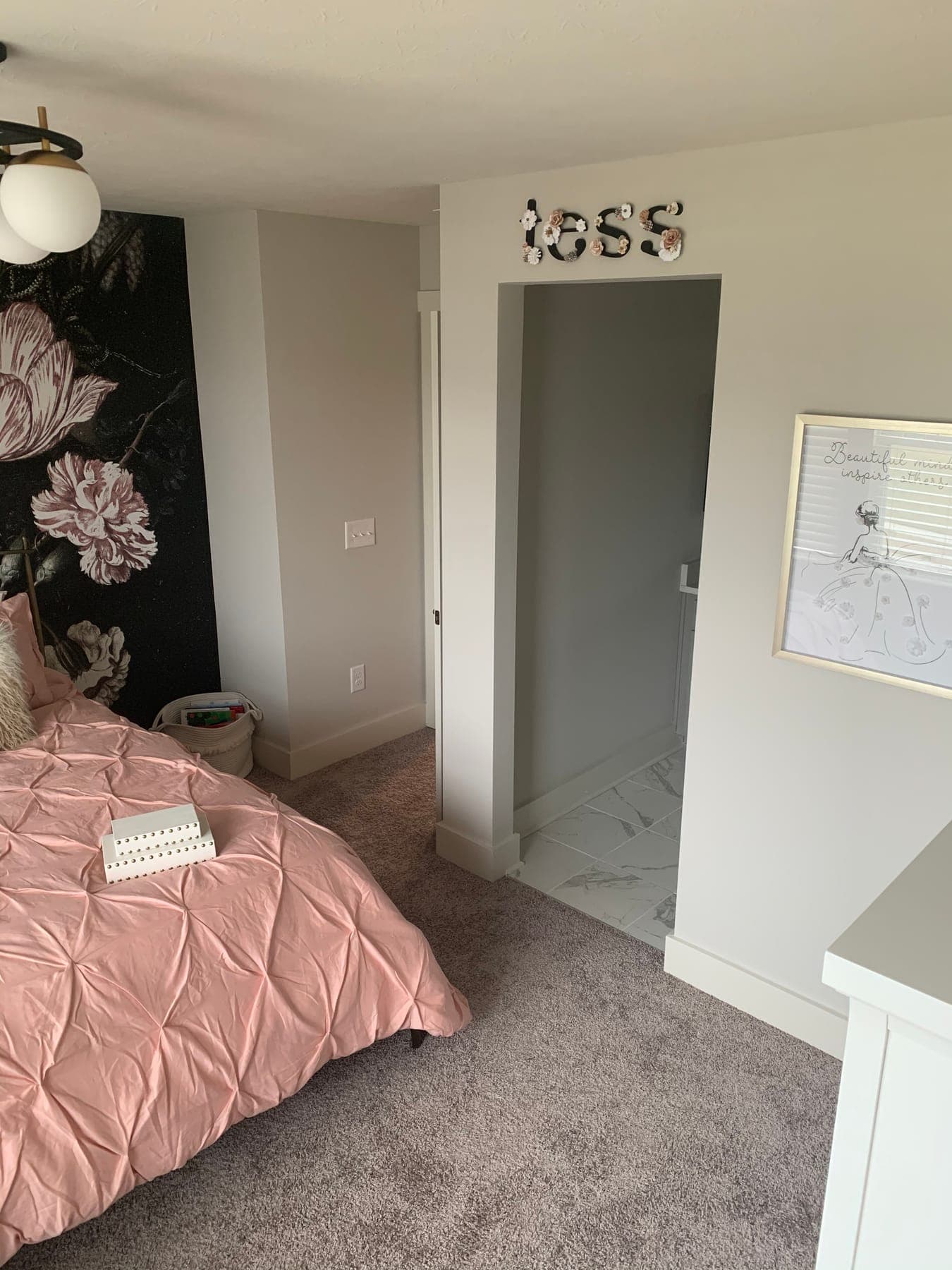 The bed and floral wall from the doorway, marquee letters spelling 'tess' above the door and a glass globe ceiling light.