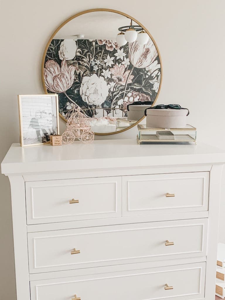 The dresser top styled with a wire carriage, a letterboard, perfume, and a box, the floral wall reflected in the mirror.