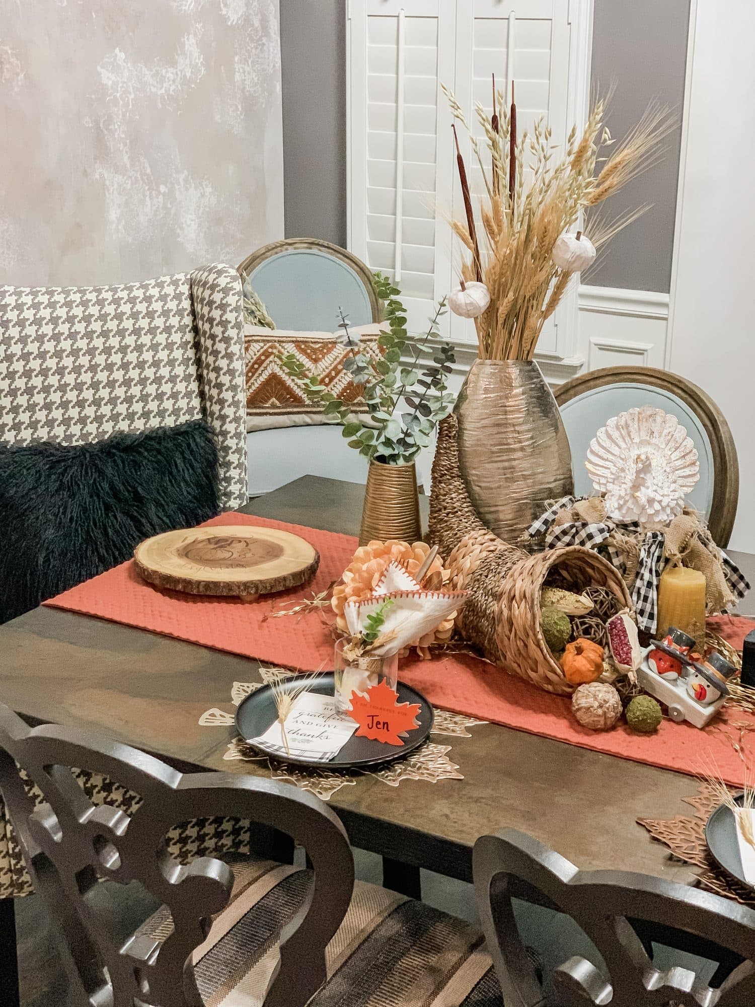 The centerpiece up close, a woven cornucopia of gourds, pinecones, and dried hydrangea over a burnt-orange runner, with a wood-slab charger and leaf place card.