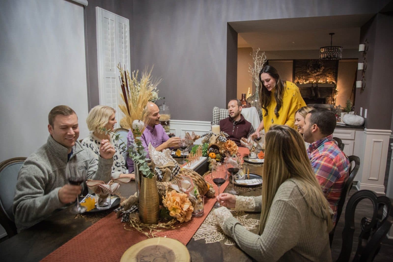 The whole table: friends around the harvest spread, Haley standing at the head in a mustard sweater, glasses up.