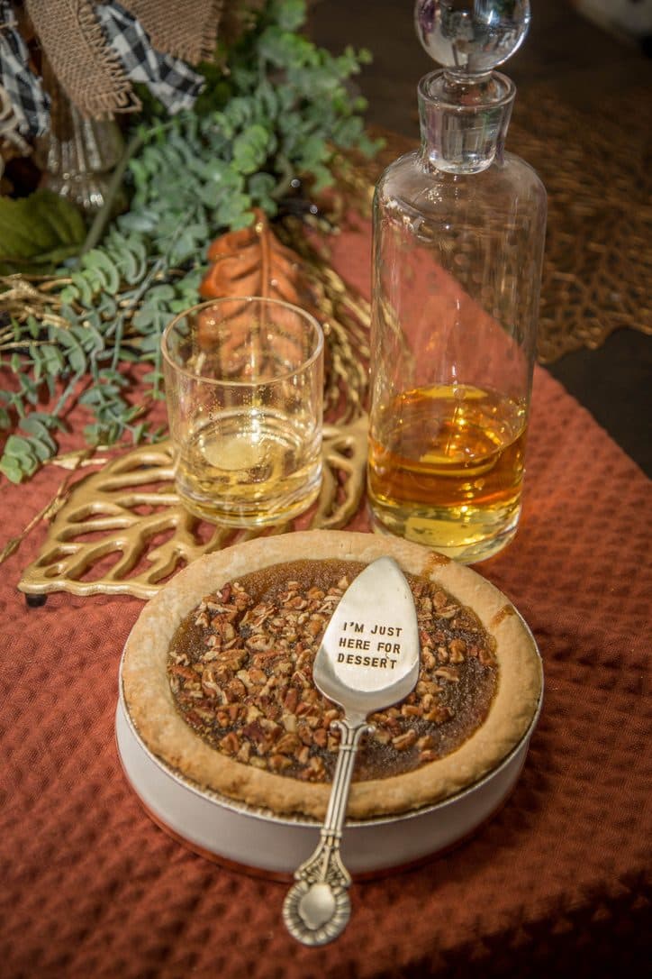 A pecan pie with an 'I'm just here for dessert' serving spoon, beside a cut-crystal decanter of amber whiskey on the orange runner.