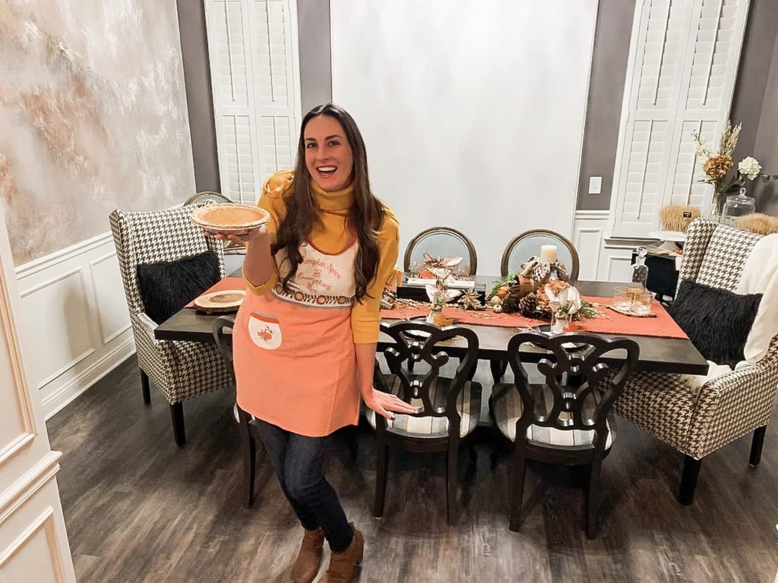 Haley in an orange apron presenting the pie to the room, houndstooth wingbacks and the styled table behind her.