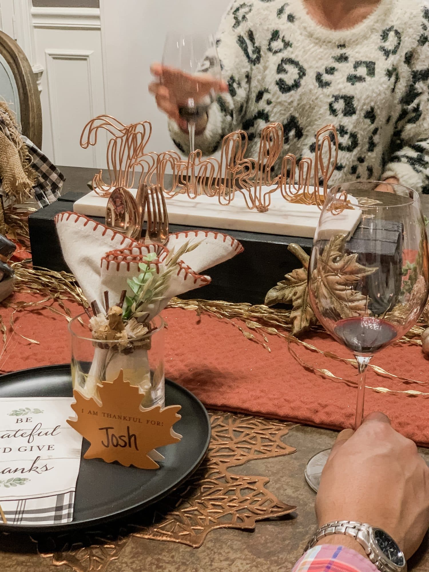 A place-setting detail, a copper wire 'Thankful' sign behind feather-wrapped flatware in a jar, a black charger with a 'Josh' leaf name card, and a copper leaf placemat on the burnt-orange runner.