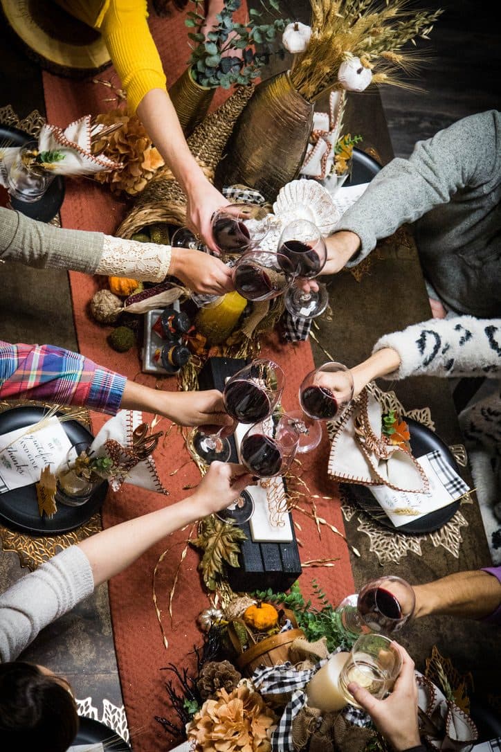 An overhead toast, hands raising red-wine glasses together over the styled centerpiece of wheat, gourds, and lace-edged napkins.